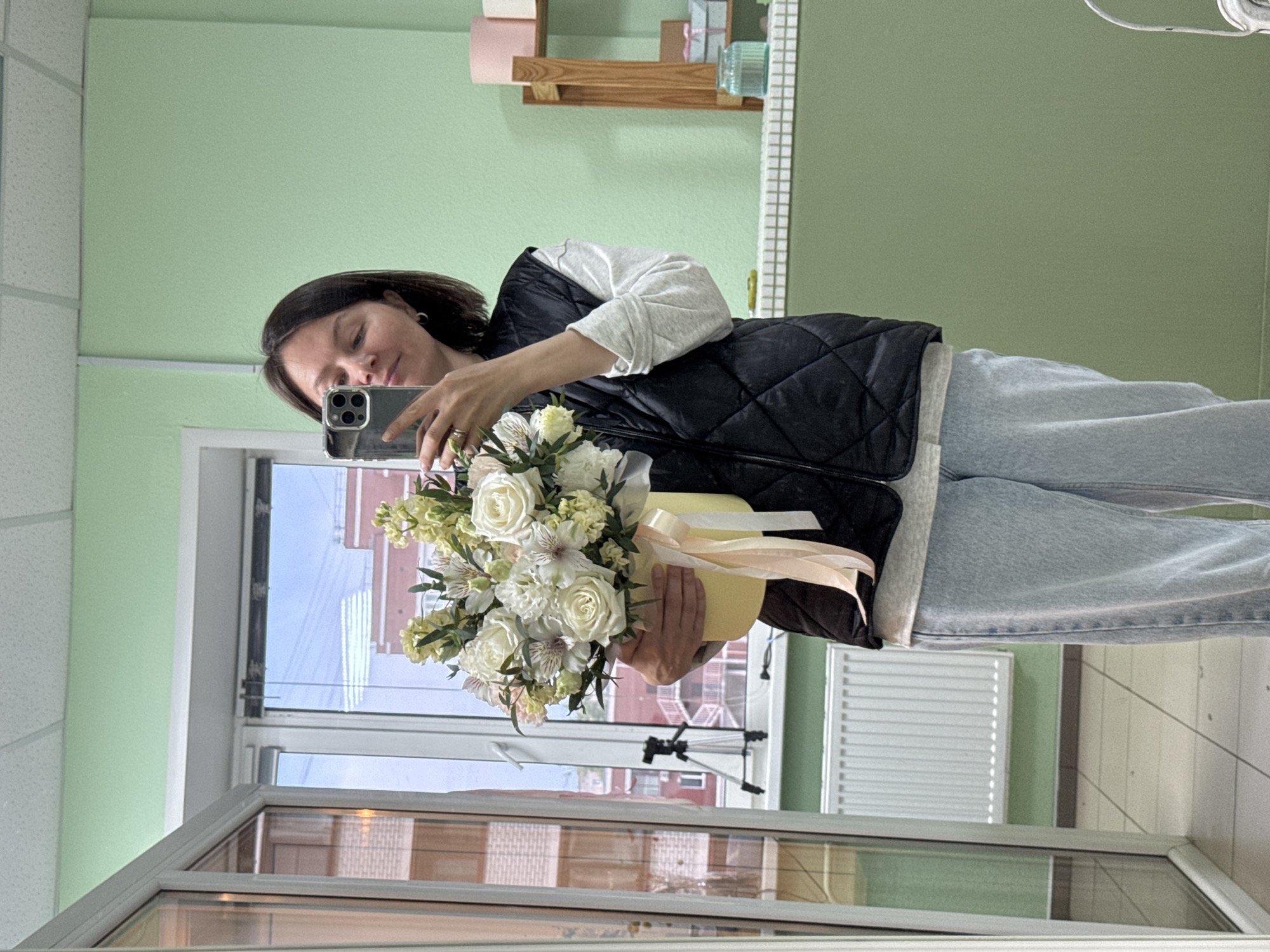 A woman is captured in a mirror selfie, holding a large bouquet of white and light yellow flowers in a pastel yellow hatbox. She has short dark brown hair, parted on the side, and is wearing a light grey long-sleeved shirt, a black quilted vest, and light blue jeans. Her right hand, adorned with rings on the ring and pinky fingers, holds a dark-colored smartphone with a prominent triple camera array, reflecting her face. A tattoo is visible on her left forearm. The flower arrangement consists predominantly of white roses, white alstroemeria, light yellow stock flowers, light pink carnations, and dark green filler foliage, with light peach ribbons tied around the hatbox.
The indoor setting features light pastel green walls and a white acoustic tile ceiling. The floor is covered with light-toned tiles. A large multi-pane window on the left provides a view of multi-story residential buildings constructed from reddish-brown brick, with an overcast sky in the background. A white radiator is positioned below the window, and a small black tripod is visible on the exterior windowsill. To the right of the woman, a light wooden shelving unit displays two pastel pink cylindrical boxes on the top shelf, and a small light blue gift box with a pink ribbon on the middle shelf. A clear glass jar with a blue-green substance rests on a white tiled counter beneath the shelves. This scene is located in Izhevsk, Russia.