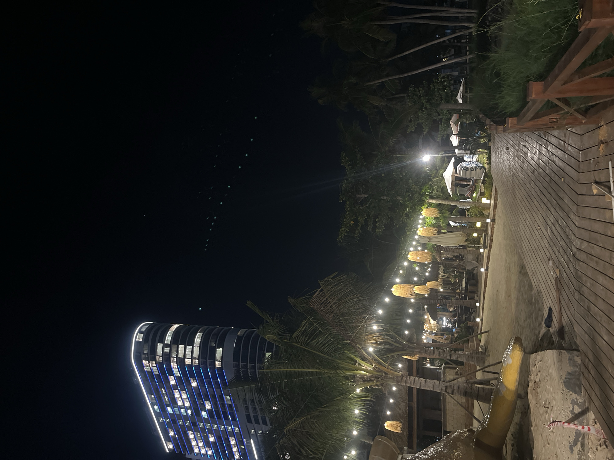 The image depicts a nighttime scene at a beachside location in Nha Trang, Vietnam, likely a resort or restaurant area. In the background, a tall, modern, curved building with blue accent lighting and illuminated windows stands against the dark sky. In the foreground, a wooden walkway extends into the scene, bordered by lush green grass on the right. The walkway leads towards a sandy area where several palm trees are present. Strung between the trees and structures are numerous string lights, illuminating the area with a warm glow. Some woven hanging lamps are also visible, adding to the ambiance. There are several thatched-roof structures and umbrellas, suggesting seating or dining areas. The sandy ground is visible, with some construction or caution tape visible on the lower left. The overall impression is a well-lit, tropical evening setting.