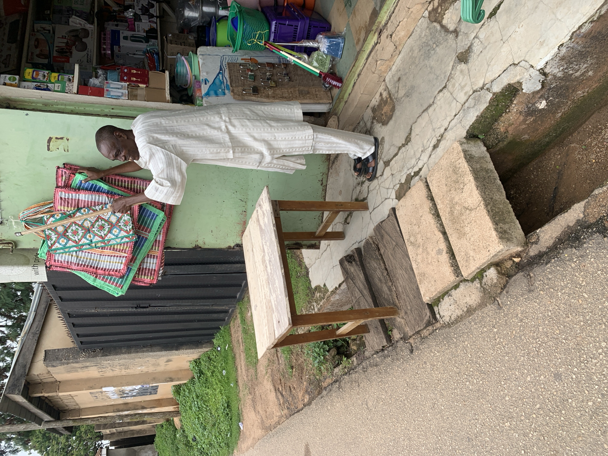 A man in a white garment is standing outside a shop, holding several colorful woven mats. The shop is decorated with the mats, which are green, red, and white with intricate patterns. The man is smiling and appears to be arranging the mats on the wall. In front of the shop, there is a wooden table and a few concrete blocks used as stepping stones. The street is paved with asphalt. The background shows a building with a black metal fence and some trees. The scene is set in Jos, Nigeria.