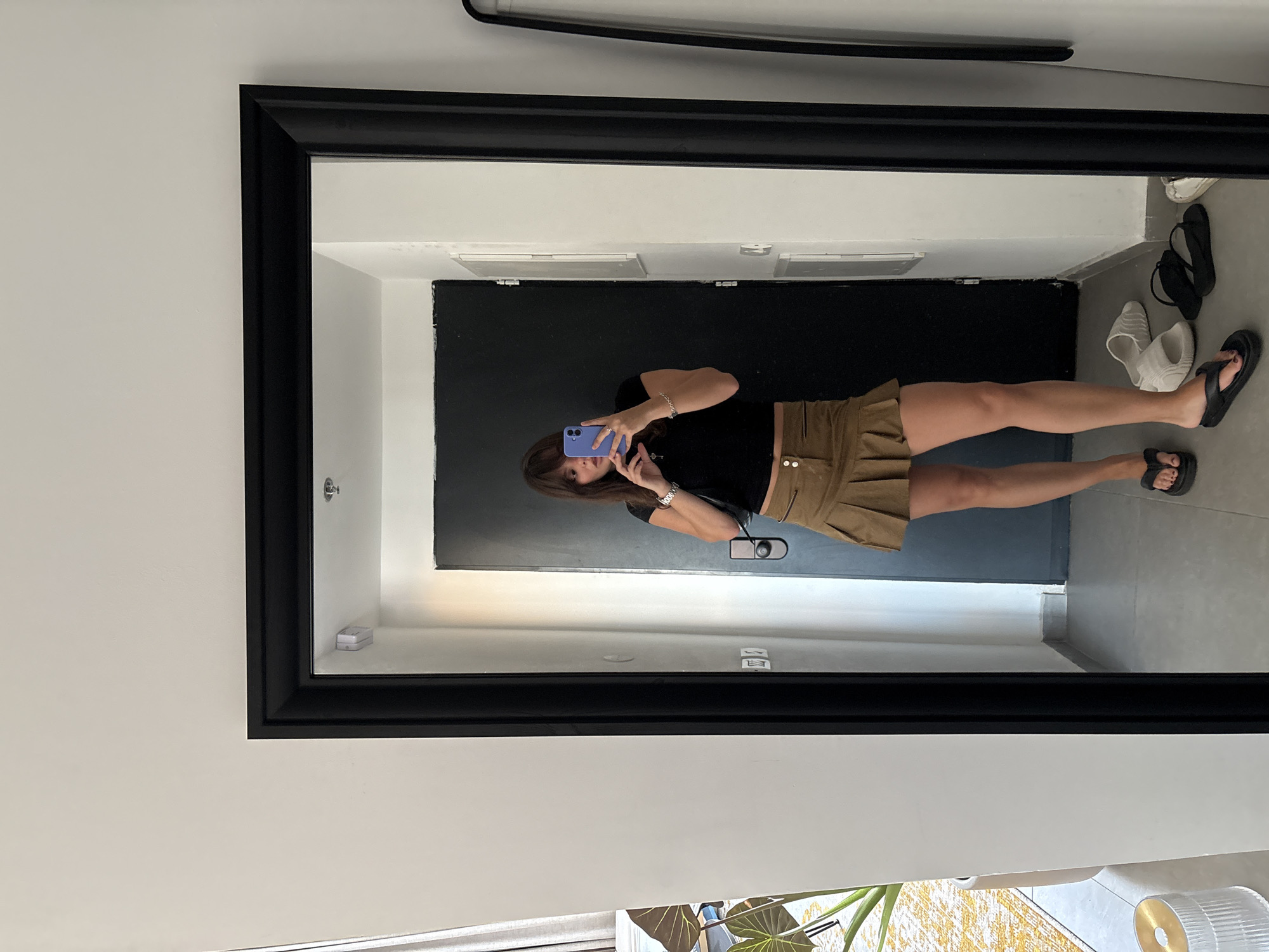 A woman, reflected in a large, black-framed mirror, takes a selfie with her phone. She is wearing a black t-shirt and a tan A-line skirt with ruffled detailing and decorative buttons. She has a silver bracelet on her left wrist and a ring on her left index finger. She is also wearing black flip-flops.
The scene reflected in the mirror shows an indoor setting. To the left of the woman's reflection, we see a doorway with a black door. To the right of the door, several pairs of footwear are visible on the floor: a pair of white slide sandals, a pair of black flip-flops, and another pair of black flip-flops. The floor appears to be tiled. The walls are white. A thin black bar runs along the top of the mirror's frame.
The photo appears to be taken indoors, possibly in a dressing room or hallway, given the presence of shoes. The lighting suggests it's daytime. The woman's posture and the selfie she is taking indicate a casual and perhaps self-aware moment.