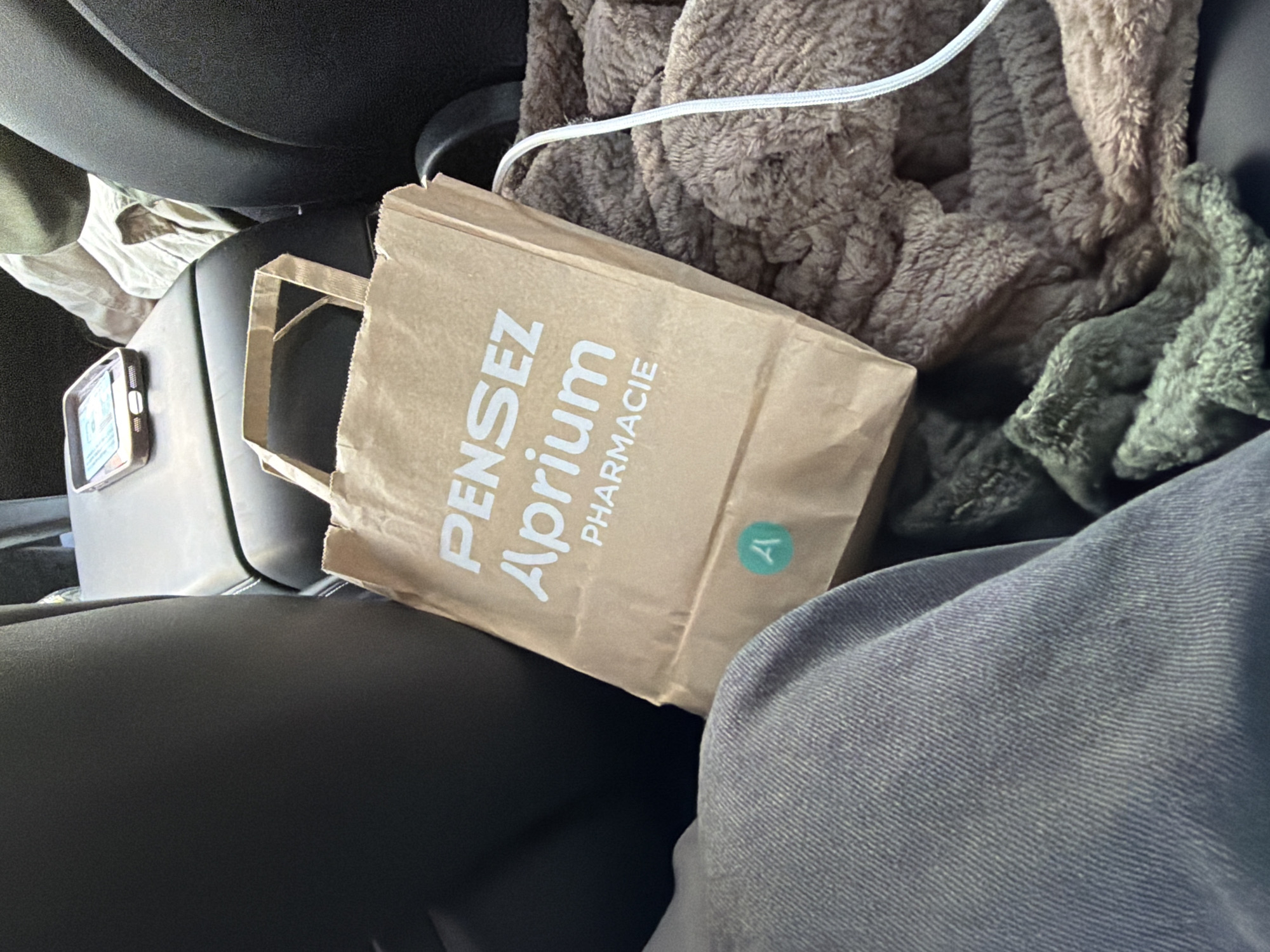 The image shows a brown paper bag with white lettering that reads "Pensez Aprium Pharmacie" placed on the center console of a vehicle. A light blue sticker with a white 'A' is affixed to the lower part of the bag. To the right of the bag, a thick, fuzzy brown blanket is visible, and a white cord extends from it. In the foreground, a person's lap covered in gray denim pants is in view. The background consists of the dark interior of a car, with portions of a car seat and the back of another seat visible. The overall scene suggests a person is inside a car, likely in the city of Ödsmål, Sweden, with a shopping bag from a pharmacy.