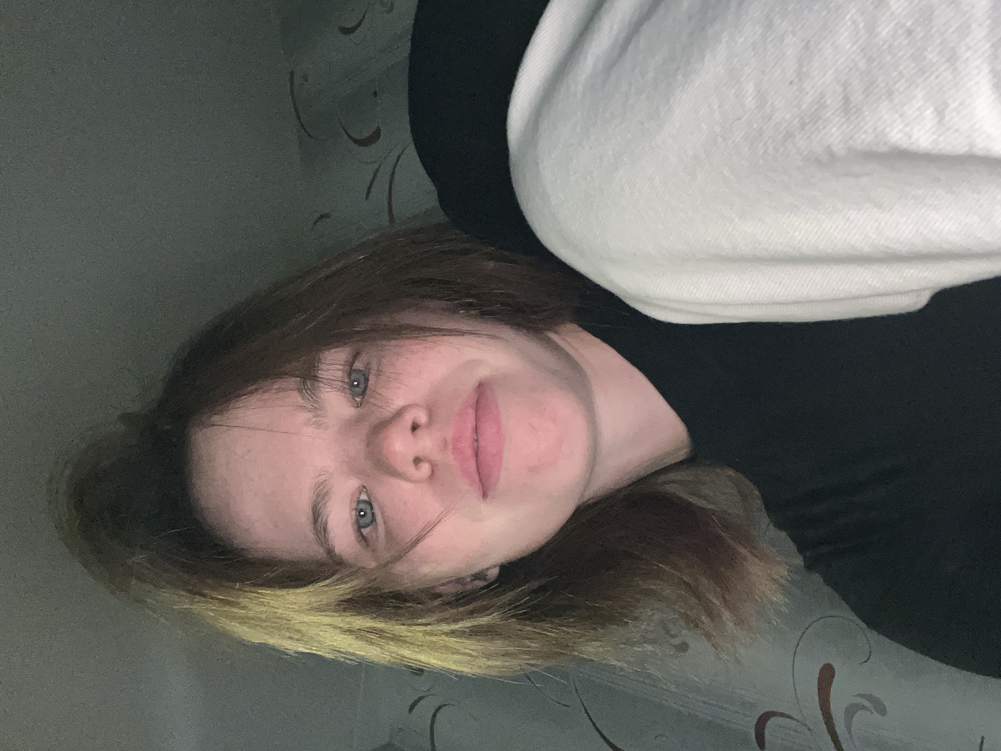 A portrait-oriented image, rotated 90 degrees clockwise, features a young person, likely female, posing for a selfie while lying down indoors. The individual has fair skin, light blue eyes, and medium-brown hair with lighter, possibly blonde, strands framing their face. Their lips are slightly parted, and they gaze directly at the camera with a neutral, calm expression.
The person is wearing a dark, possibly black, long-sleeved top, partially obscured by a white, textured hooded garment or blanket that covers their shoulder and upper chest. The background consists of a wall adorned with a subtly patterned wallpaper, featuring ornate, swirling designs in shades of grey and reddish-brown. The lighting appears soft, indicating an indoor setting, possibly a room like a bedroom or living area.
There are no discernible activities beyond posing for the photograph. The time of day is not explicitly clear but suggests artificial or dim natural lighting. The overall mood is relaxed and intimate. No text is visible in the image. The location is Chaikovsky, Russia.