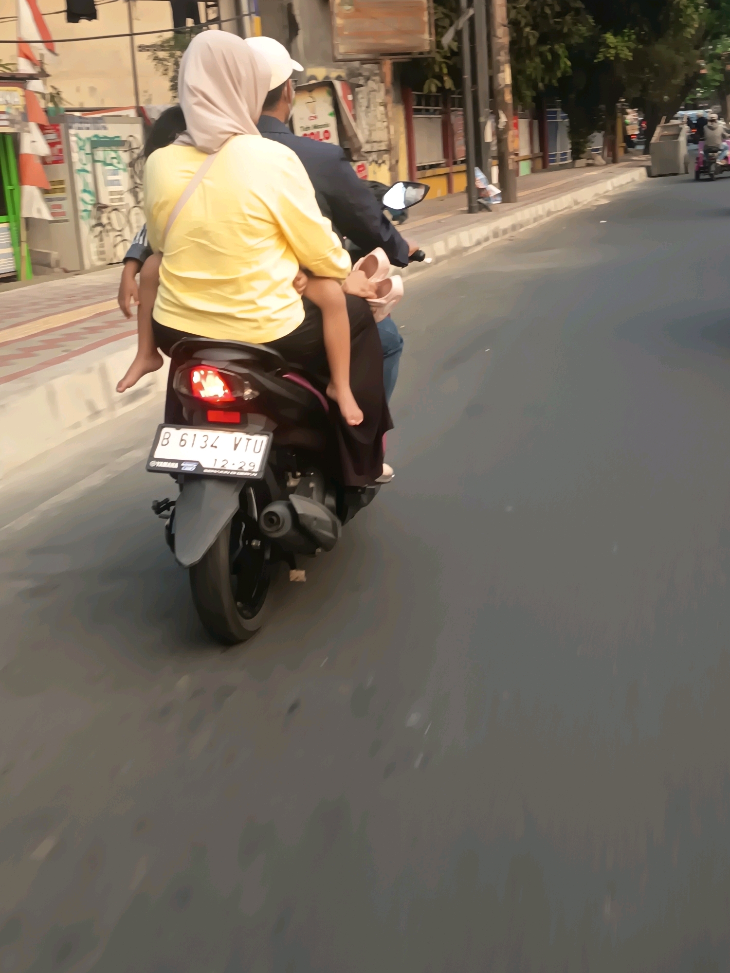 This is a medium-shot, eye-level, full-color photograph taken from behind a motorcycle on a paved road in Tangerang, Indonesia. The motorcycle, a dark-colored scooter, is moving forward, indicated by the motion blur of the road and surrounding environment. On the back of the motorcycle, a woman wearing a yellow long-sleeved shirt and a light-colored headscarf is holding a small child. The child, partially visible, is wearing a light-colored shirt and has bare feet. The woman is wearing dark pants, and the child is also wearing dark pants. A beige strap is visible across the woman's right shoulder. The motorcycle has a white license plate that reads "B 6134 VTU" with "12-29" below it, and a Yamaha logo. The scene is lit by natural daylight. To the left of the road, a sidewalk with red and white striped poles and some graffiti is visible. The background shows buildings, trees, and other vehicles, suggesting an urban environment. The overall impression is of a typical street scene in Indonesia.