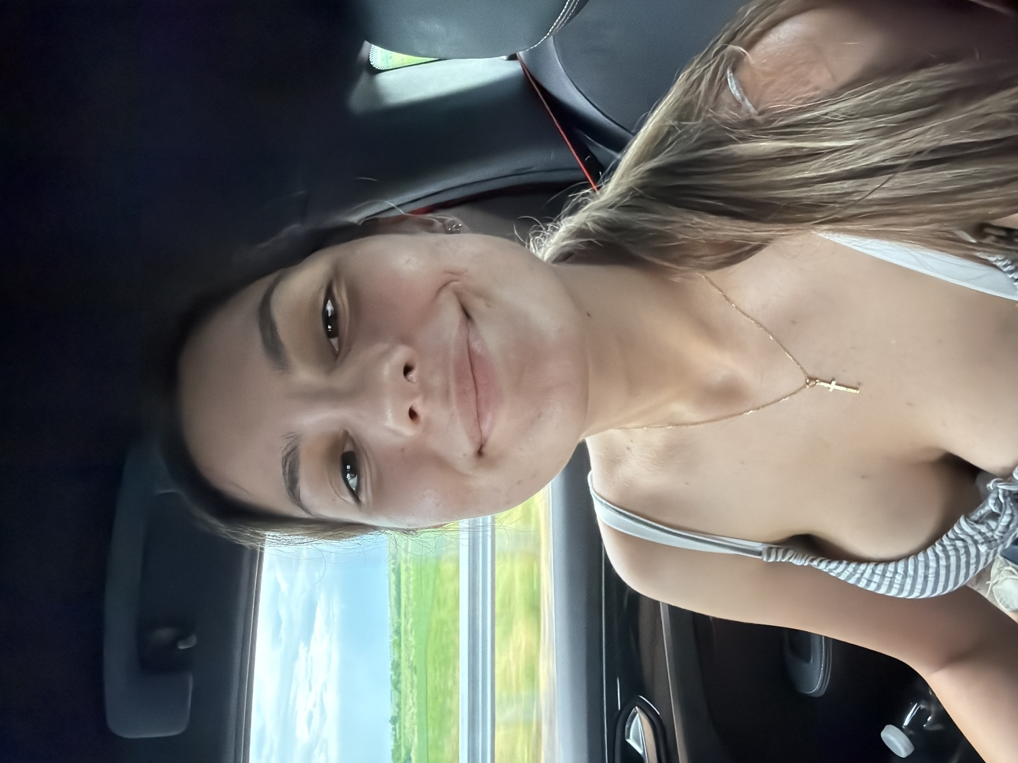 A young woman with long, light-brown hair is in the passenger seat of a car, possibly moving based on the blurred background visible through the window. She's wearing a striped top and a gold necklace with a cross pendant. She has fair skin and appears to be smiling subtly. The car's interior is visible, including the black seats and part of the door panel. Outside the window, there's a green field or grassy area under a blue and white sky. There is no location data available to indicate this is Chokoba, Bulgaria.
