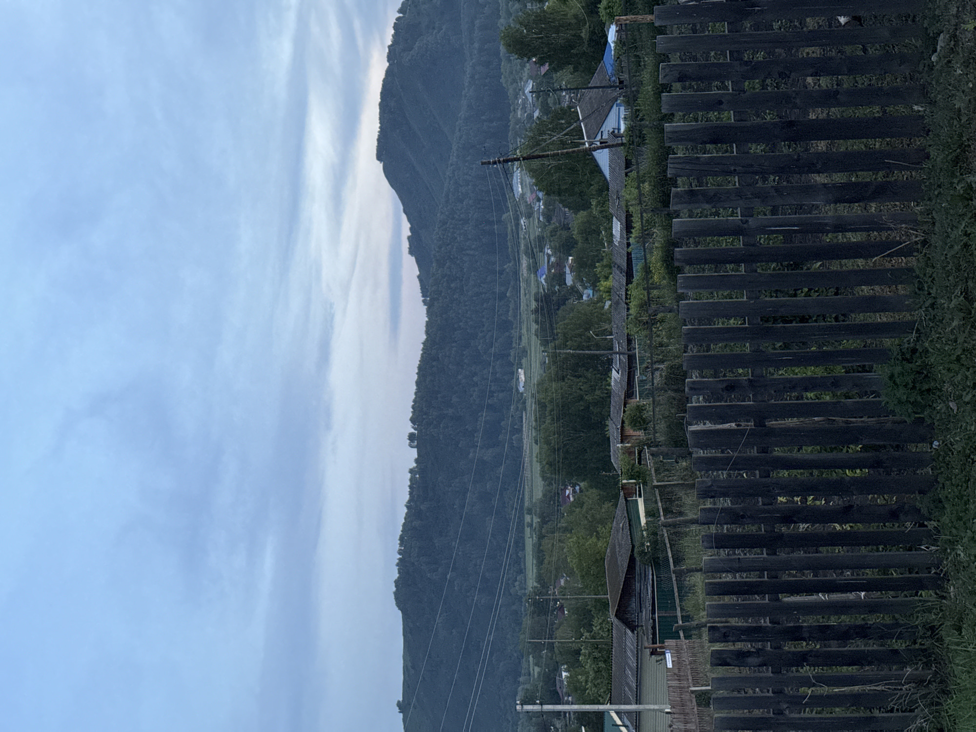 The image depicts a rural village scene at dusk, likely in Soloneshnoye, Russia. In the foreground, a weathered, dark wooden fence runs horizontally across the frame. Behind the fence, several modest, rustic houses with corrugated metal roofs are visible, interspersed with trees. Beyond the village, a large, densely forested hill dominates the middle ground, with power lines stretching across its slope. The sky above is a soft blue, with wispy clouds suggesting the transition from day to night. The overall atmosphere is serene and quiet, characteristic of a rural settlement. There are no people or animals visible in the frame.