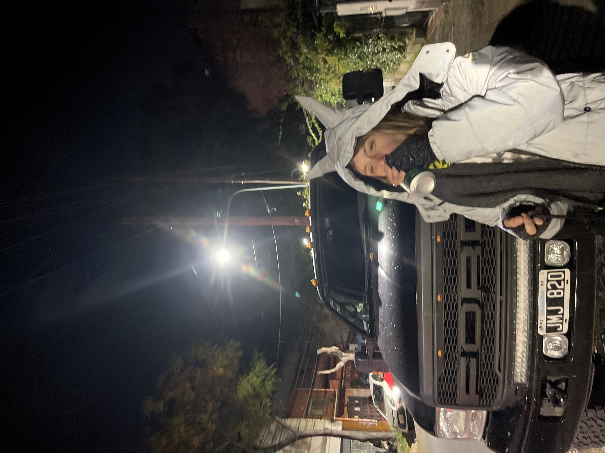 The image depicts a woman in a gray hooded jacket holding a can, standing next to a black Ford truck with license plate "JMJ 820". The location appears to be a street at night, with streetlights visible. Buildings and trees are also visible in the background. The scene is possibly in Olivos, Argentina, based on the license plate mentioning "Argentina".