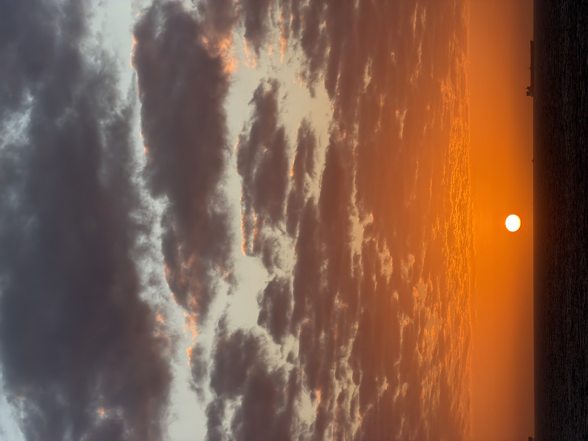 The image captures a sunset over water, likely a sea or large lake, at an unknown location. The sun is a bright, circular orb near the horizon on the right. A dark, indistinct landmass or structure is visible on the far right, possibly a coastline or ship. The sky transitions from a bright orange near the horizon to darker, cooler tones on the left. The clouds are varied in texture, with dense, dark formations on the left and lighter, wispy clouds illuminated by the sunset near the center and right. The scene lacks specific identifying features to pinpoint the exact location.