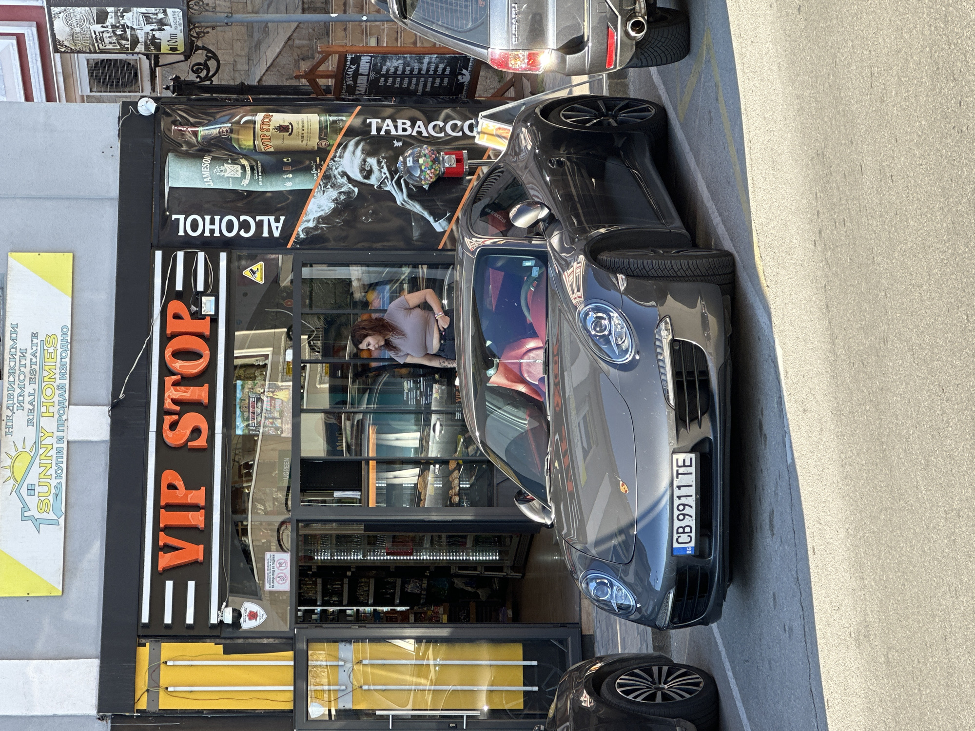 Here's a detailed description of the image:
**General Overview**
The image depicts the storefront of a "VIP STOP" establishment, likely a convenience store or a small shop selling alcohol and tobacco, in Veliko Tarnovo, Bulgaria. A silver Porsche is parked directly in front of the store. The weather appears sunny.
**Specific Elements**
* **Storefront:** The storefront has a sign that reads "VIP STOP" in large, illuminated orange letters. Above that, the sign reads "ALCOHOL" and "TABACCO" with an image of a man smoking. There is an advertisement for Jameson Irish Whiskey. A sticker in the store window says UGREEN. The store itself seems to be stocked with various goods, visible through the windows.
* **Cars:** A gray Porsche, bearing a Bulgarian license plate "CB 9911 TE", is parked in front of the store. Part of a black SUV is seen parked behind the Porsche.
* **Person:** A woman is standing inside the store, appearing to look at her phone.
* **Signage:** The sign for Sunny Homes and advertisements are visible on the building next to the store.
* **Street:** The street is paved. Yellow lines indicating parking spaces.
**Location (Inferred from Context)**
The presence of Bulgarian text and the license plate indicate the location is in Bulgaria, specifically Veliko Tarnovo.