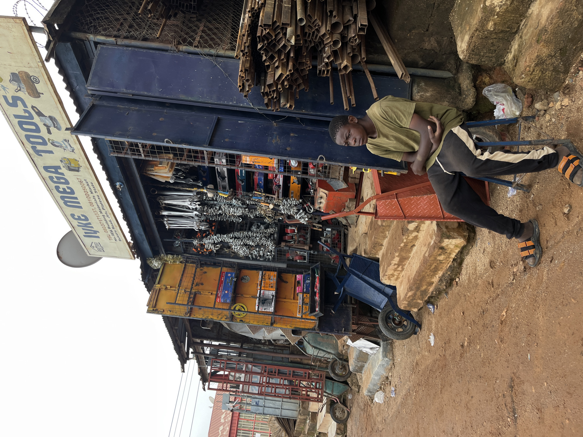 The image captures a street scene in Gura Top, Nigeria, focusing on a stall selling tools and building materials. In the foreground, a young boy in a green t-shirt and black and white striped track pants sits on a blue stool. He is wearing black and orange striped sandals. His arms are crossed, and he is looking to his left. The stall itself is a colorful assortment of goods, with various tools, wrenches, and chains hanging on display. Boxes of tools are stacked on shelves within the stall. To the right of the stall, a large pile of metal pipes is stacked. Various carts and trolleys, including a blue hand truck, are also present, suggesting a busy marketplace environment. The ground is dirt and appears to be a street or pathway. In the background, more stalls and buildings are visible, hinting at a densely populated commercial area. The sky is overcast.