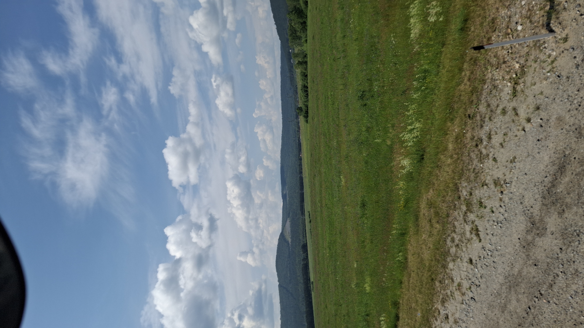 A wide-angle, eye-level shot captures a rural landscape near Shelekhov, Russia, under a partly cloudy sky. The foreground is a gravel road transitioning into a vast, green field dotted with small wildflowers. In the middle ground, a dense forest of dark green trees stretches across the horizon, backed by rolling hills of a lighter green. The sky is a vibrant blue, streaked with wispy white cirrus clouds and interspersed with larger, puffy cumulus clouds. The overall scene is serene and natural.