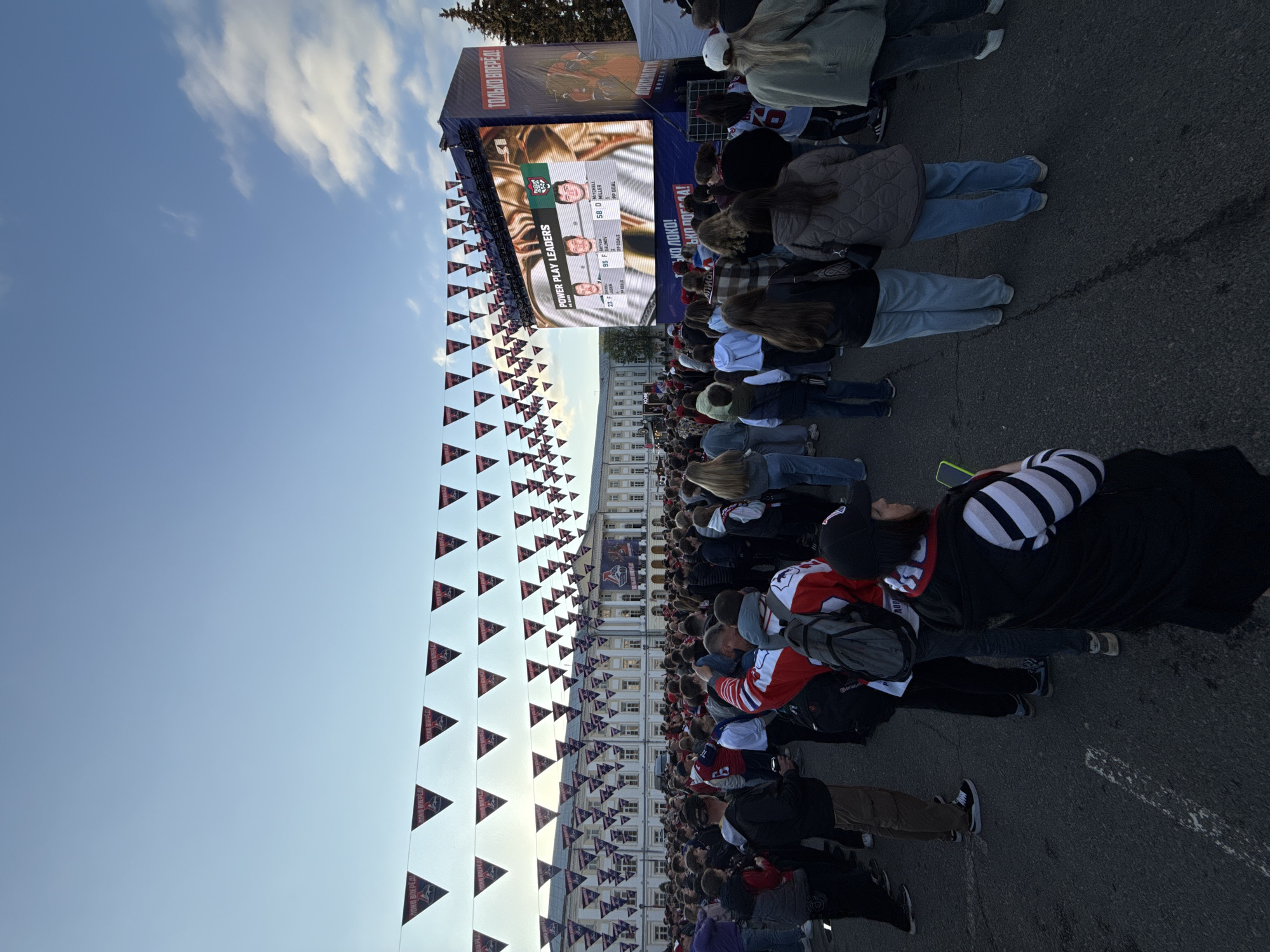 A low-angle, wide shot of a large crowd of people gathered in an open square in what is likely Unknown, Unknown. In the background, a long, light-colored building with many windows is visible. In the center, a large screen displays "POWER PLAY LEADERS" with portraits of three individuals, situated on a raised platform or stage. Banners displaying text reading "ЛОКО ВПЕРЕД!" and "ТОЛЬКО ВПЕРЕД!" are strung above the crowd. The sky above is partially cloudy, with blue patches visible. The crowd appears to be watching or engaged with the content on the screen.