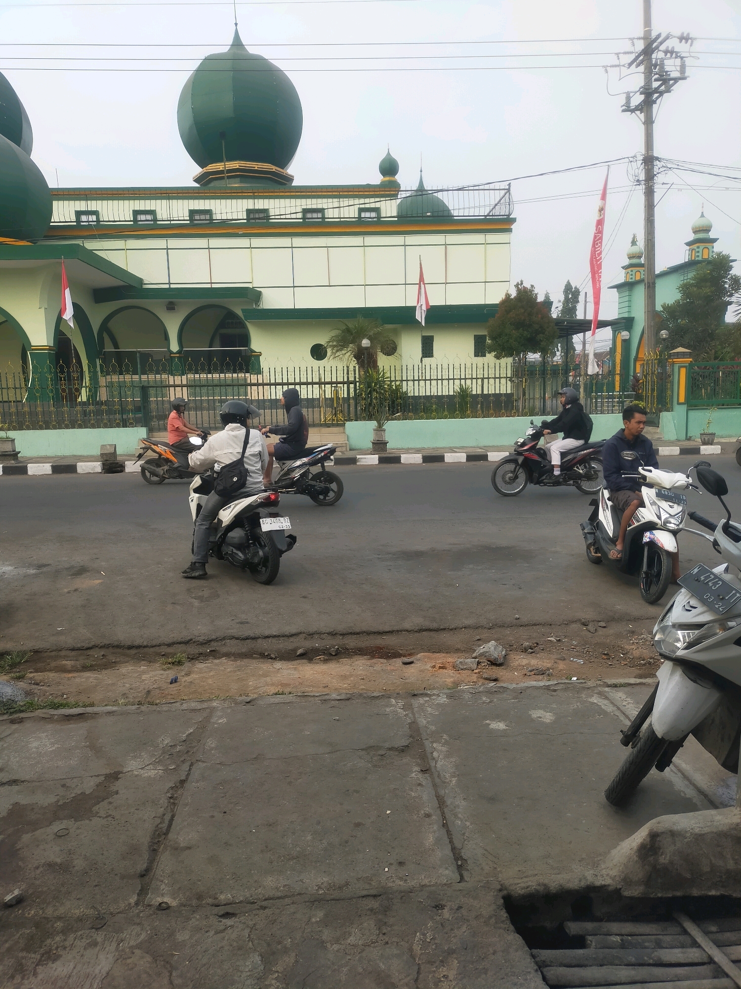 The image depicts a street scene in Bululawang, Indonesia, with a mosque in the background. A large, green-domed mosque with white and green accents dominates the upper left of the frame. In front of the mosque, a road runs horizontally across the image. Several people are riding motorcycles on the road. In the foreground, a white scooter is parked on the right side, and another white scooter is partially visible. The road appears to be paved with asphalt and has a curb on the left. The area is somewhat overcast, with utility poles and wires visible against the sky. Several red and white Indonesian flags are also displayed on poles in front of the mosque.