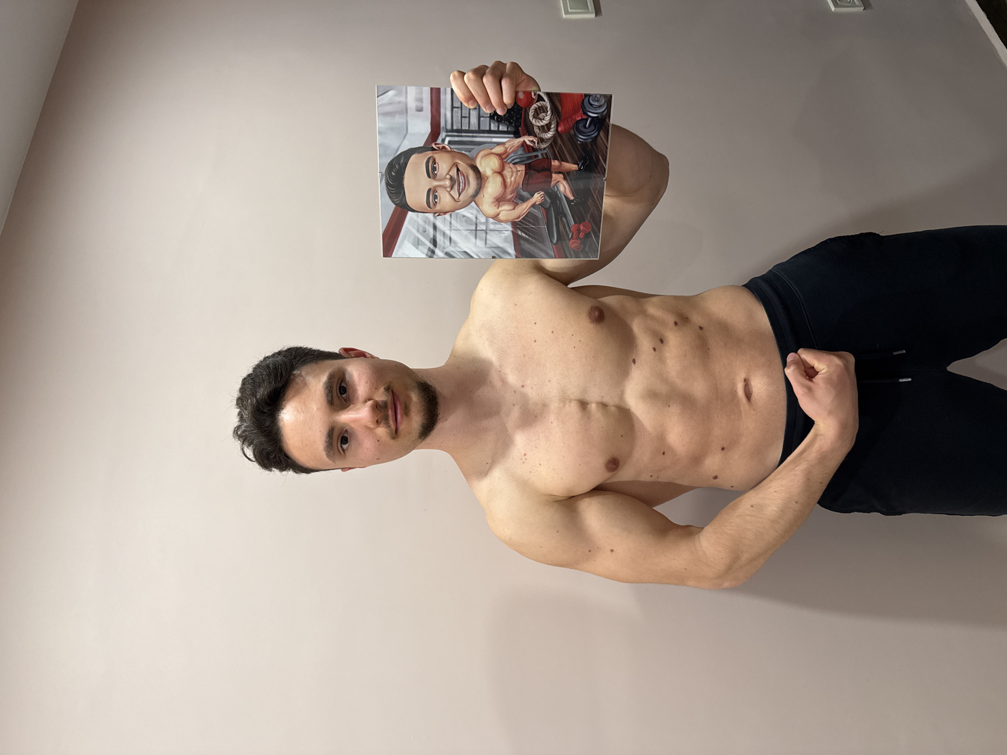 The media file displays a young, bare-chested man with an athletic physique standing upright in front of a plain, light-colored wall; the image itself is rotated 90 degrees clockwise. The man has short dark hair and a neatly trimmed short beard or stubble, and he looks directly at the camera with a neutral expression. His toned torso shows visible abdominal muscles and several small moles or freckles. He is wearing dark pants.
With his right hand, the man holds up a small, rectangular print of a personalized caricature. This cartoon depiction features a similar-looking man with an exaggerated muscular physique and a wide, smiling face. The caricature's background suggests a gym environment, with details like dumbbells, battle ropes, and a weight bench visible. The cartoon figure is also bare-chested and appears to be wearing shorts. The man's left hand is resting near his waist, clenched into a loose fist. The setting is indoors, likely a room in a home, indicated by the simple wall and a light switch or outlet visible in the upper background. There is no visible text in the image.
