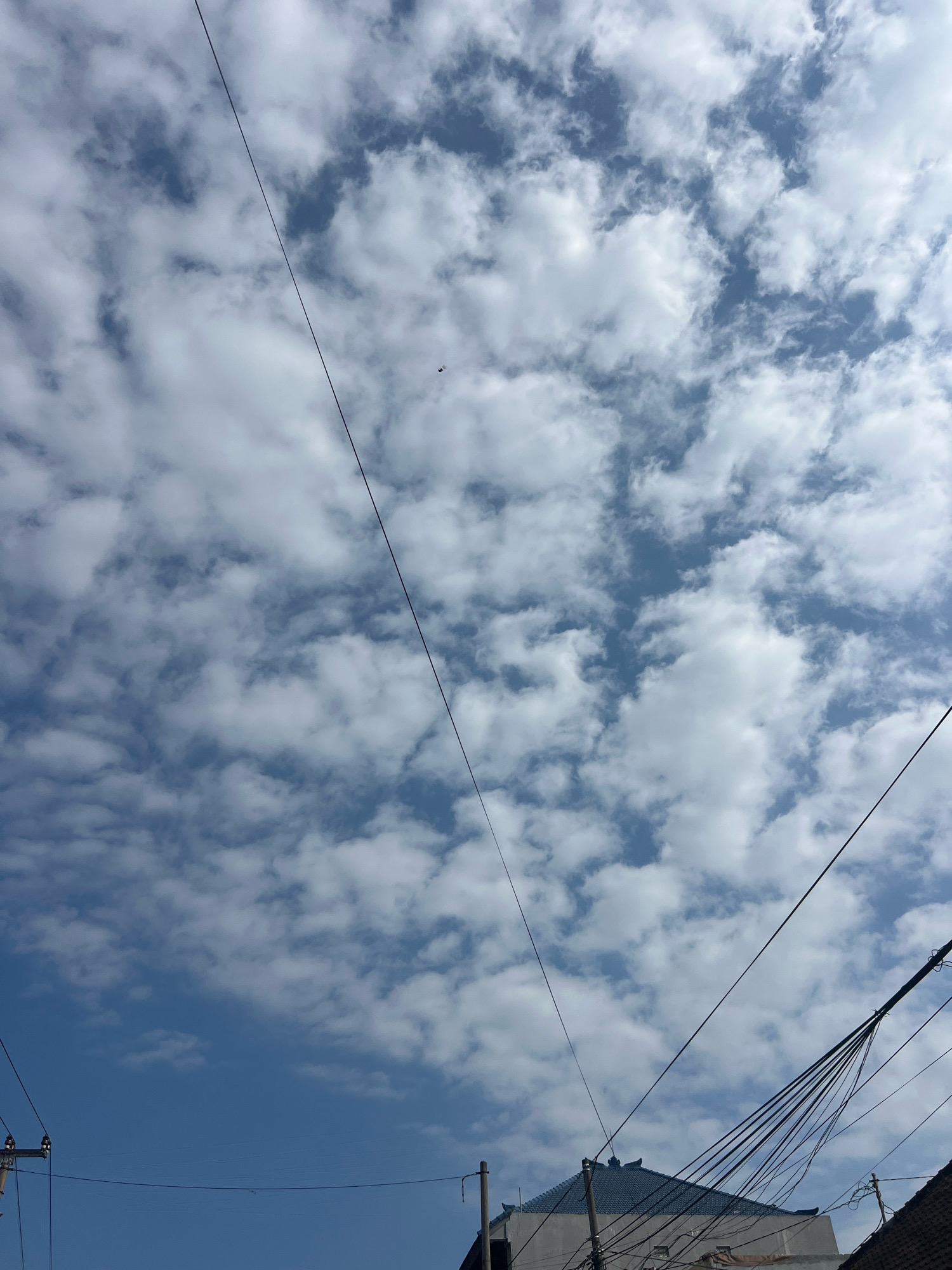 This is a medium shot, angled upwards, of a cloudy blue sky with several electrical wires strung across the frame. In the lower portion of the image, the rooftops of a few buildings are visible, some with dark tiled roofs and others with lighter colored facades. The sky is filled with scattered white cumulus clouds, with patches of clear blue sky showing through. The electrical wires are black and appear to be suspended from poles, one of which is visible on the left side of the frame. The scene appears to be in an urban or semi-urban area, possibly Denpasar, Indonesia, given the architectural style of the buildings and the prevalence of overhead electrical wires. The image is taken during daylight hours.