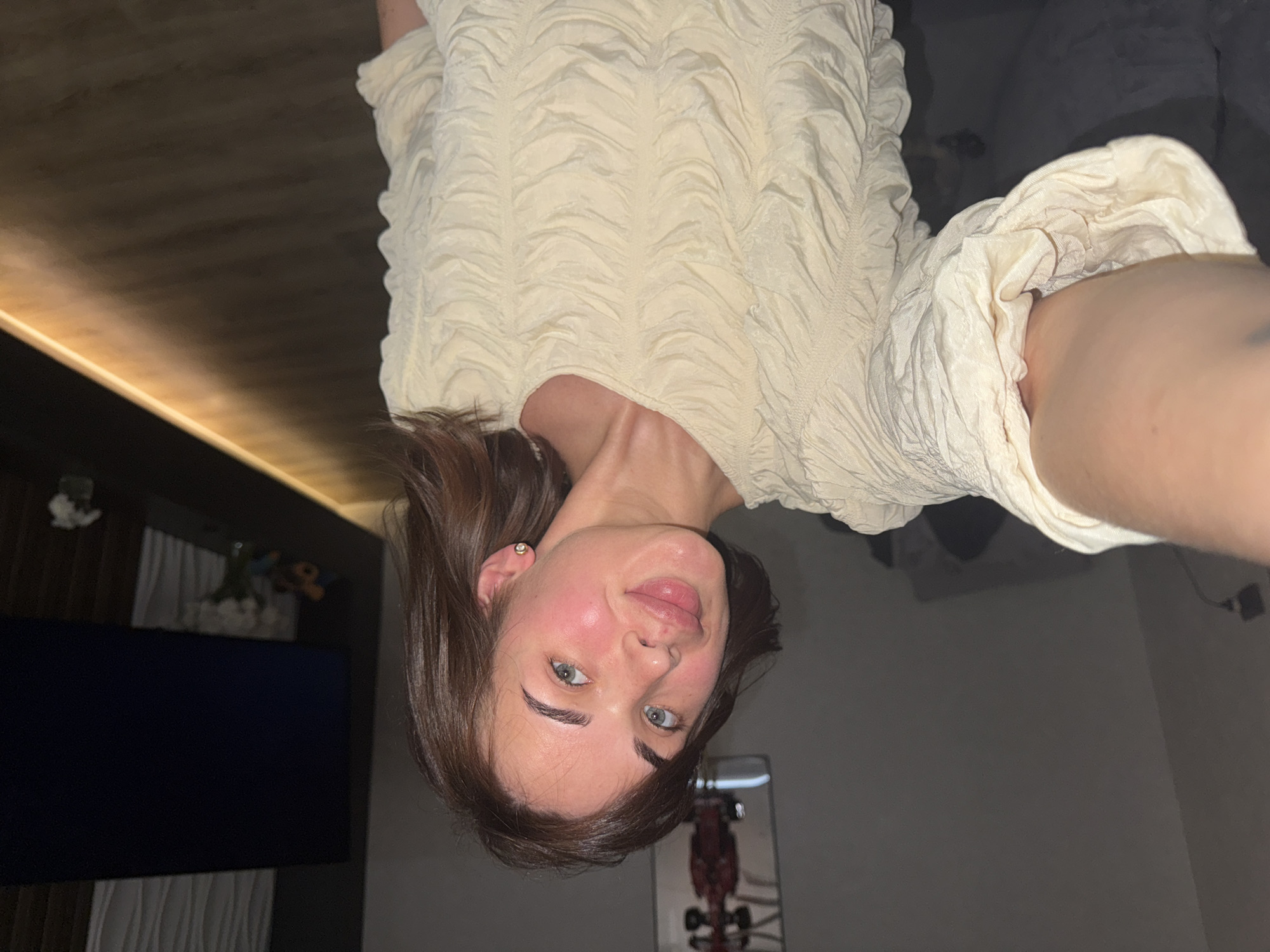 A woman with shoulder-length brunette hair and blue eyes is facing the camera. She is wearing a light-colored, textured top. She appears indoors in Лобаново, Russia. Behind her, a television is visible on the left, and a display case is partially visible on the lower right. The room has a dark ceiling with a strip light and gray walls.