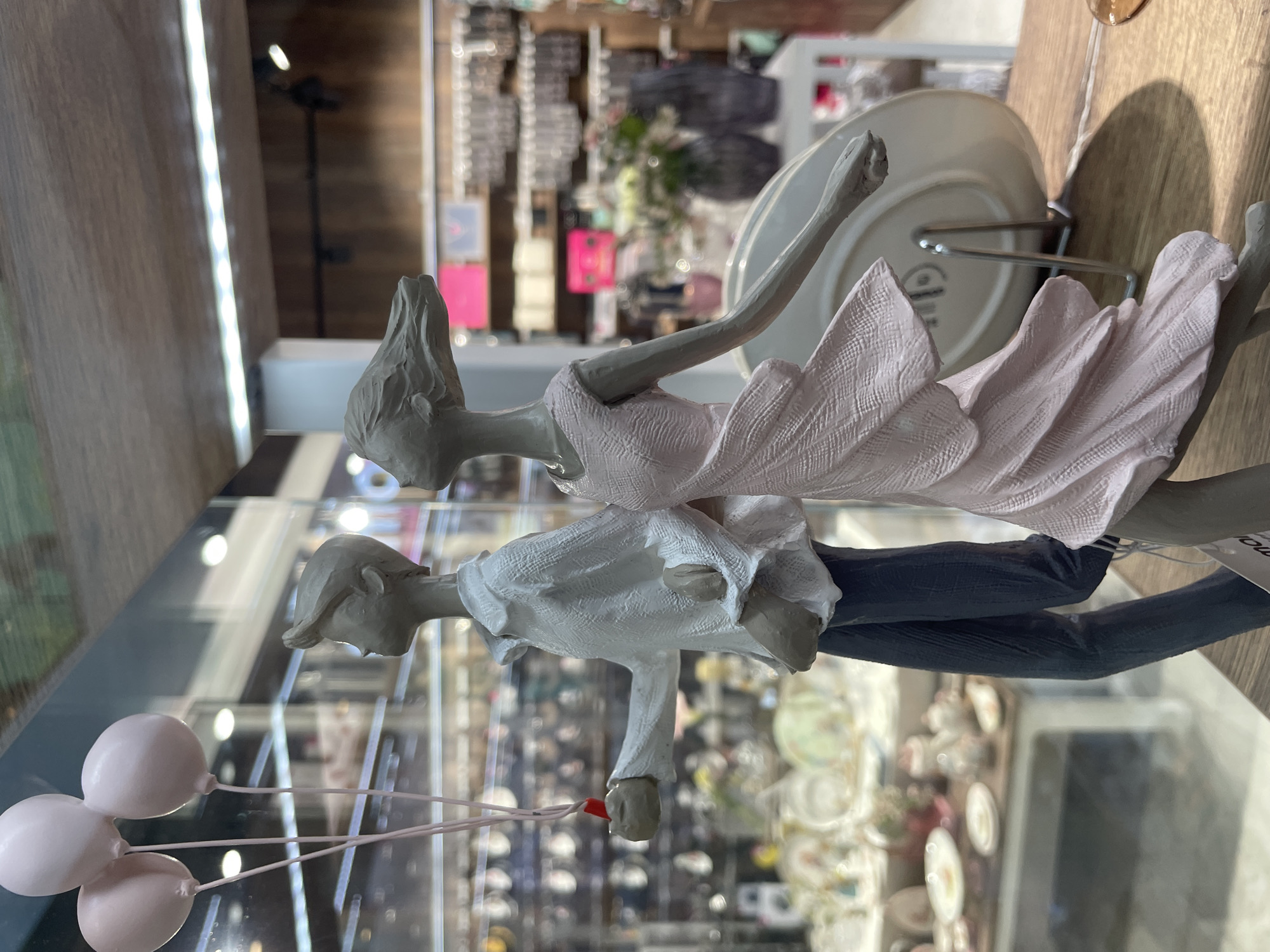 The image shows a ceramic figurine of a couple in love. The man is wearing a white shirt and dark pants and holding three light pink balloons. The woman is wearing a pink dress and has her arm raised. The figurine is displayed on a glass shelf. Behind the figurine is a blurry background of shelves stocked with various items. Based on the available information, it is impossible to confirm that the location is in Kommunarka, Russia.