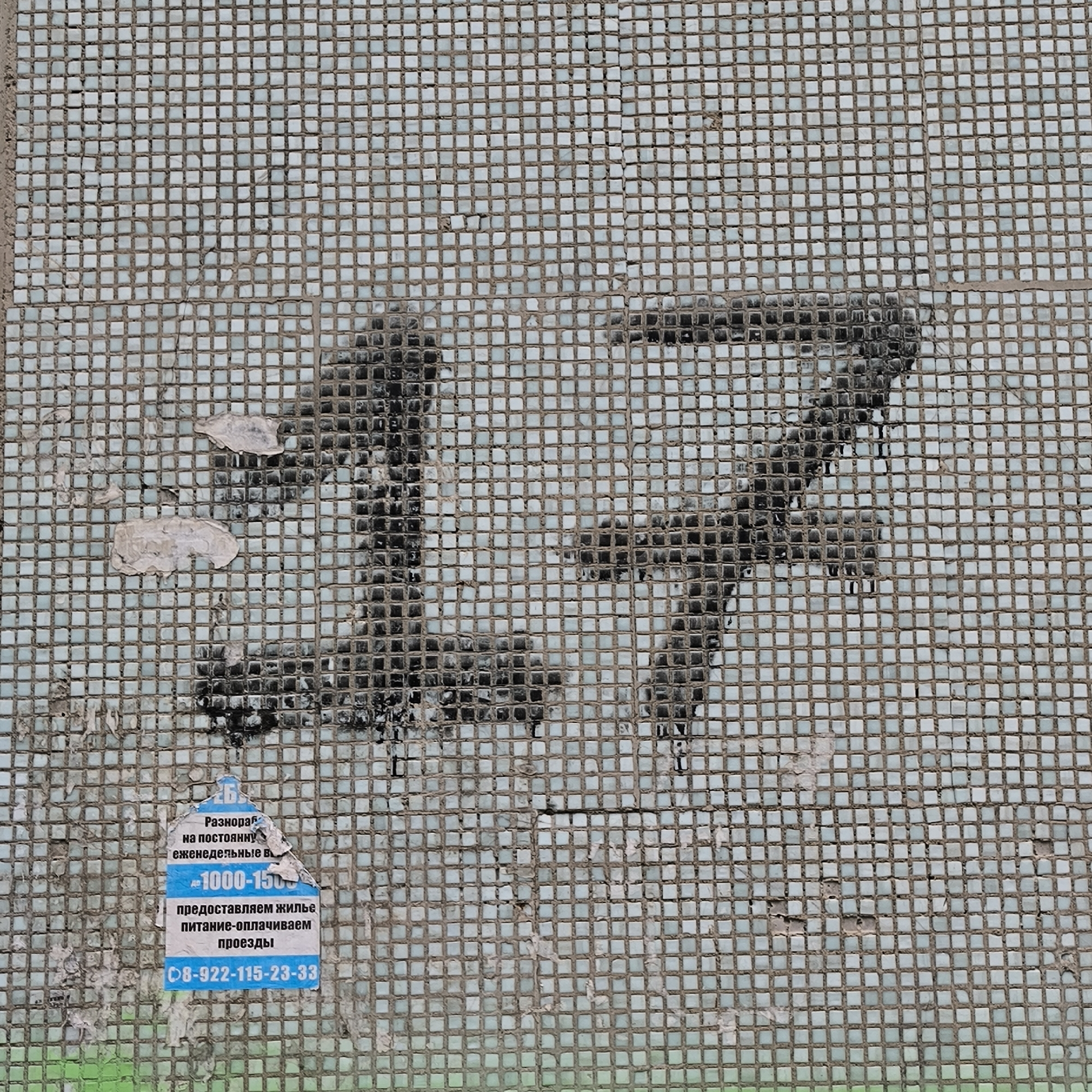 The image is a close-up shot of a mosaic tiled wall, likely on the exterior of a building. The mosaic tiles are small, square, and light blue or white, with visible grout lines forming a grid pattern. The wall appears weathered, with some tiles missing and patches of grime or paint obscuring parts of the mosaic.
Prominently displayed on the wall are the large, spray-painted numerals "17". The numbers are rendered in black, with a rough, uneven texture, suggesting they were applied with a stencil or freehand. The paint is partially faded and streaked, further indicating age and exposure to the elements.
In the lower left quadrant of the image, a torn advertisement poster is affixed to the mosaic wall. The poster is predominantly blue and white, with text in Russian. The visible text includes "Разнорабочий на постоянную", "еженедельные в", "1000-1500", "предоставляем жилье", "питание-оплачиваем", "проезды", and a phone number "08-922-115-23-33". This advertisement appears to be for a job offering, possibly for general labor, with stated weekly wages and benefits.
The overall environment suggests an urban setting, possibly in a residential or public area. Based on the advertisement text in Russian and the context of similar images found online, this mosaic wall with the number "17" is located at **2b Komsomolskaya Street, Miass, Russia**. It is a common feature of Soviet-era panel buildings and serves as an unofficial building or entrance number.