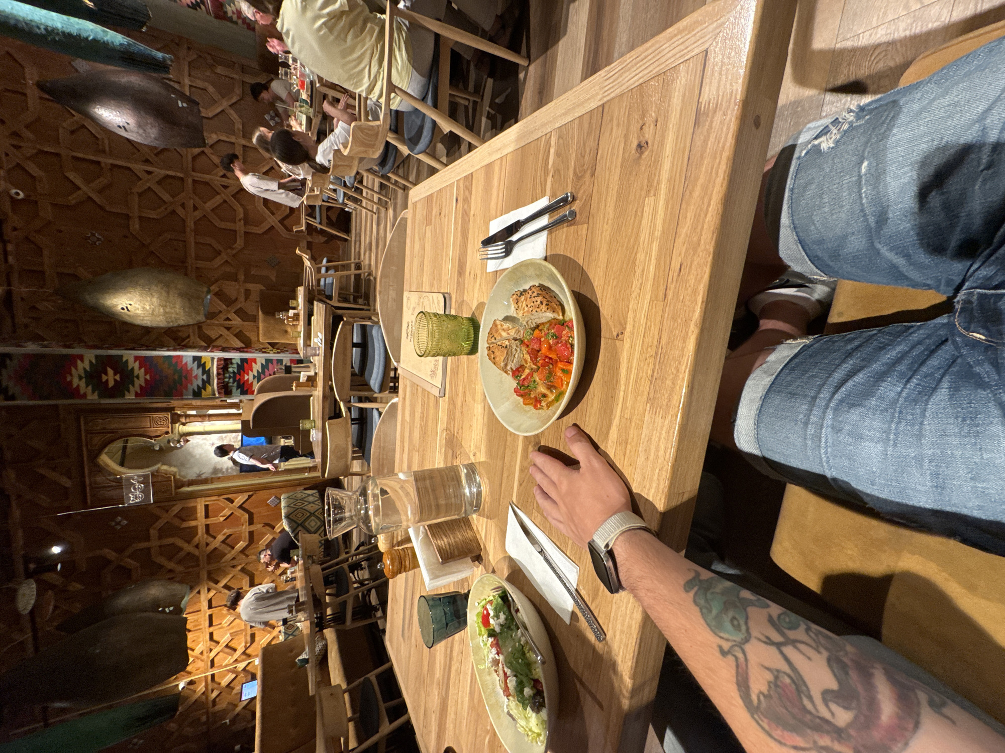 The image depicts the interior of a restaurant. In the foreground, a wooden table is laden with food and drinks, including a salad, a plate of chicken and vegetables, a glass of water, and a green glass. A person's tattooed arm and legs in denim jeans are visible, suggesting they are seated at the table. In the background, other diners are seated at tables, and the restaurant features ornate wooden wall paneling and decorative lighting fixtures. The overall ambiance suggests a modern, ethnic-inspired dining establishment. There is no specific indication of the location being in Saratov, Russia.