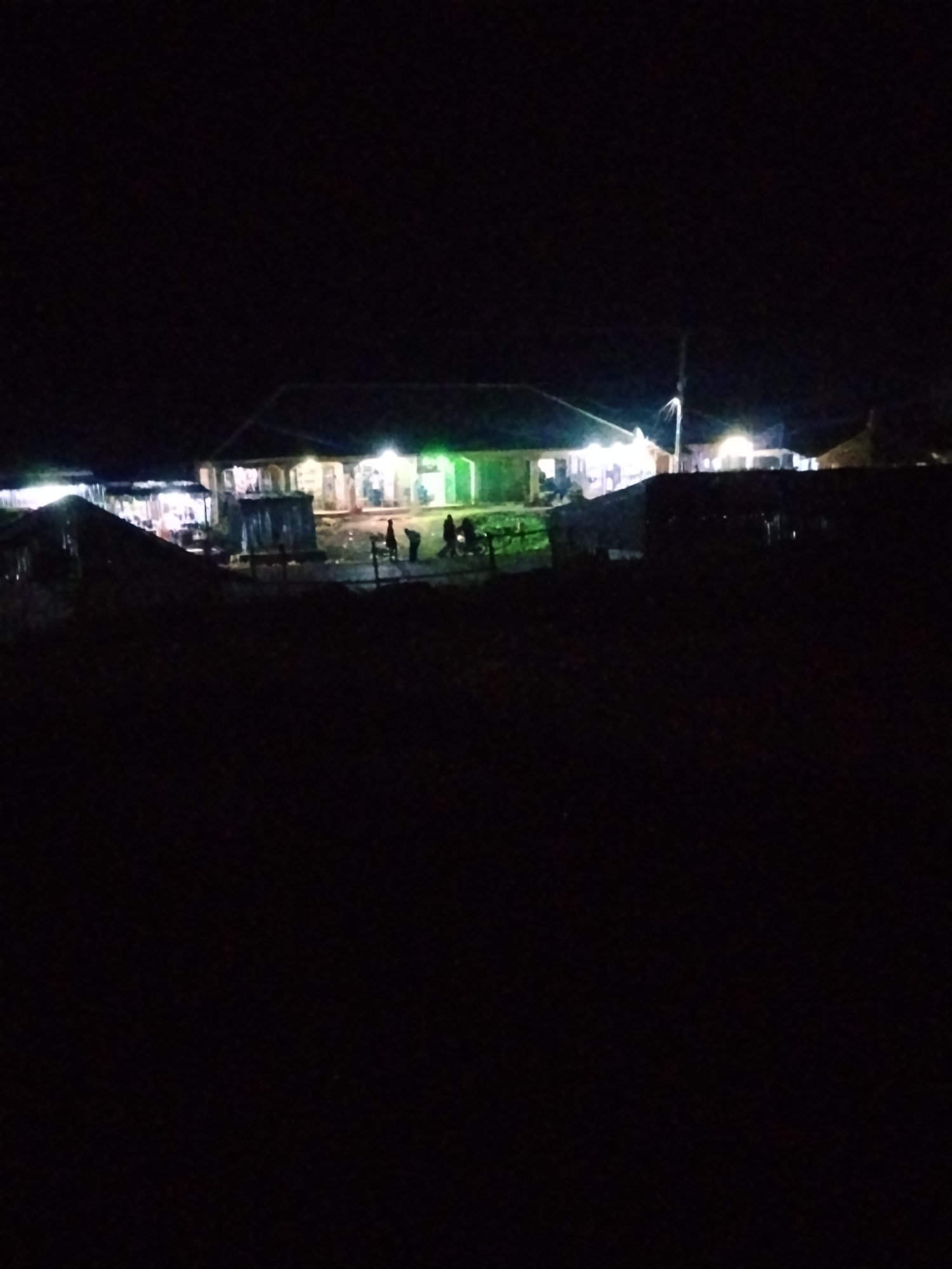 A dark, outdoor night scene features a long, low-lying building illuminated by multiple lights, suggesting it is a commercial establishment or residence. The building has a dark roof and lighter-colored walls with visible windows and doorways. Several figures, appearing as silhouettes due to the low light, are positioned in front of and near the building, possibly engaged in activities such as gathering or walking. Some appear to be standing around or moving. The foreground is obscured by deep shadow, rendering any details in that area indiscernible. A faint, vertical pole or structure is visible to the right of the building. The overall impression is one of a dimly lit, possibly bustling area at night in a location that appears to be rural or on the outskirts of a town or city. The context suggests this could be a market or a gathering place.