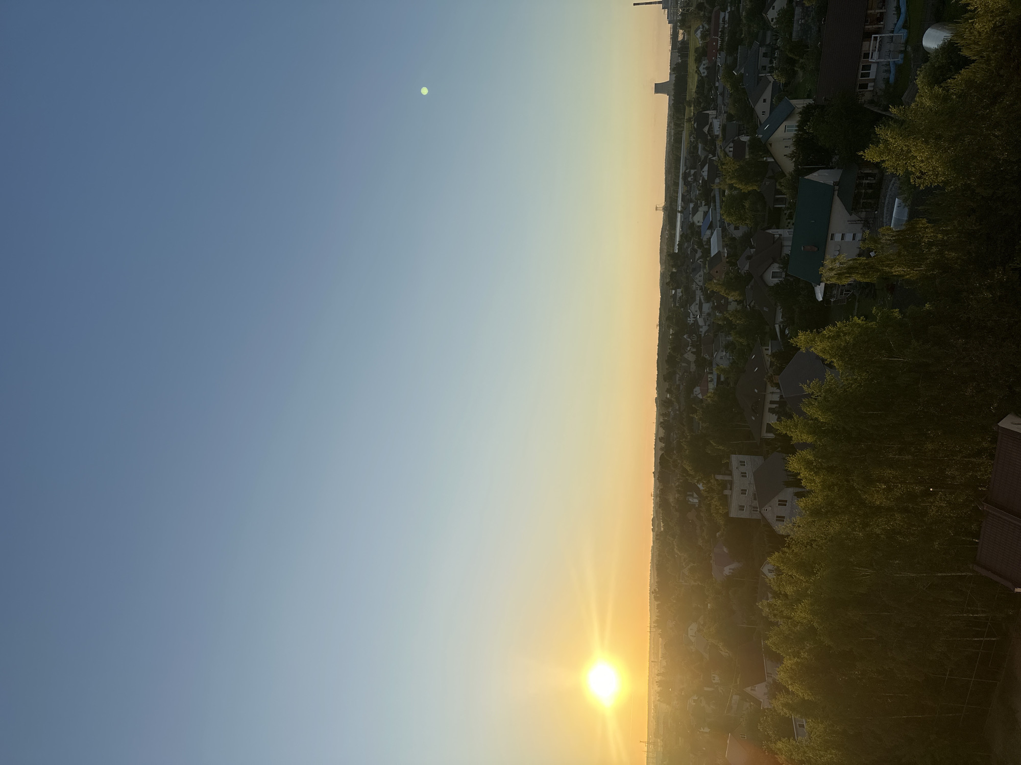 The image presents an aerial view of a suburban landscape during sunset, likely in the Шчомысліцкі сельскі Савет, Belarus. The sun, a bright orb casting a warm orange glow, is low on the horizon to the left, illuminating the sky with shades of yellow and orange that transition to a clear blue overhead. A faint, pale green circle, possibly the moon or a lens flare artifact, is visible in the upper right portion of the sky.
Below the sky, a dense collection of residential buildings with pitched roofs, varying in color and material, are nestled amongst lush green trees. Many of the homes appear to be detached houses with surrounding gardens and foliage. In the distance, a large industrial cooling tower and a chimney stack are discernible, suggesting the presence of a power plant or factory. The foreground is dominated by the canopy of trees, with some rooftops peeking through. The overall scene depicts a tranquil, rural-like setting within a developed area, bathed in the soft light of dusk. No people are visible in the image.