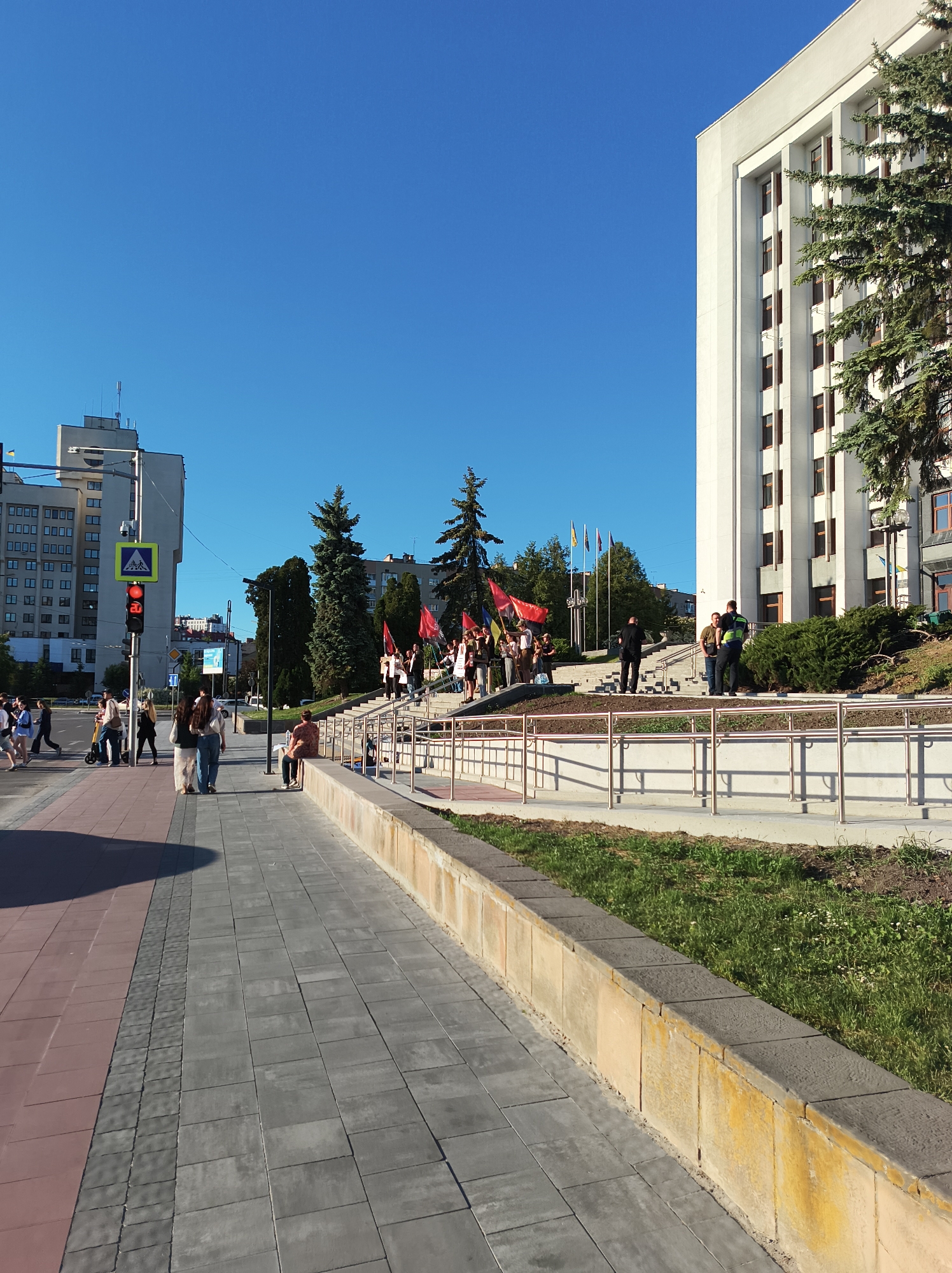 The image shows a street scene in Ternopil, Ukraine. On the left side of the frame, pedestrians are crossing at a crosswalk with a lit traffic signal. A group of people holding red flags are gathered in front of a white building with a row of national flags. Two men in uniform appear to be interacting with the group. The environment includes trees, grass, sidewalks, and a clear blue sky.