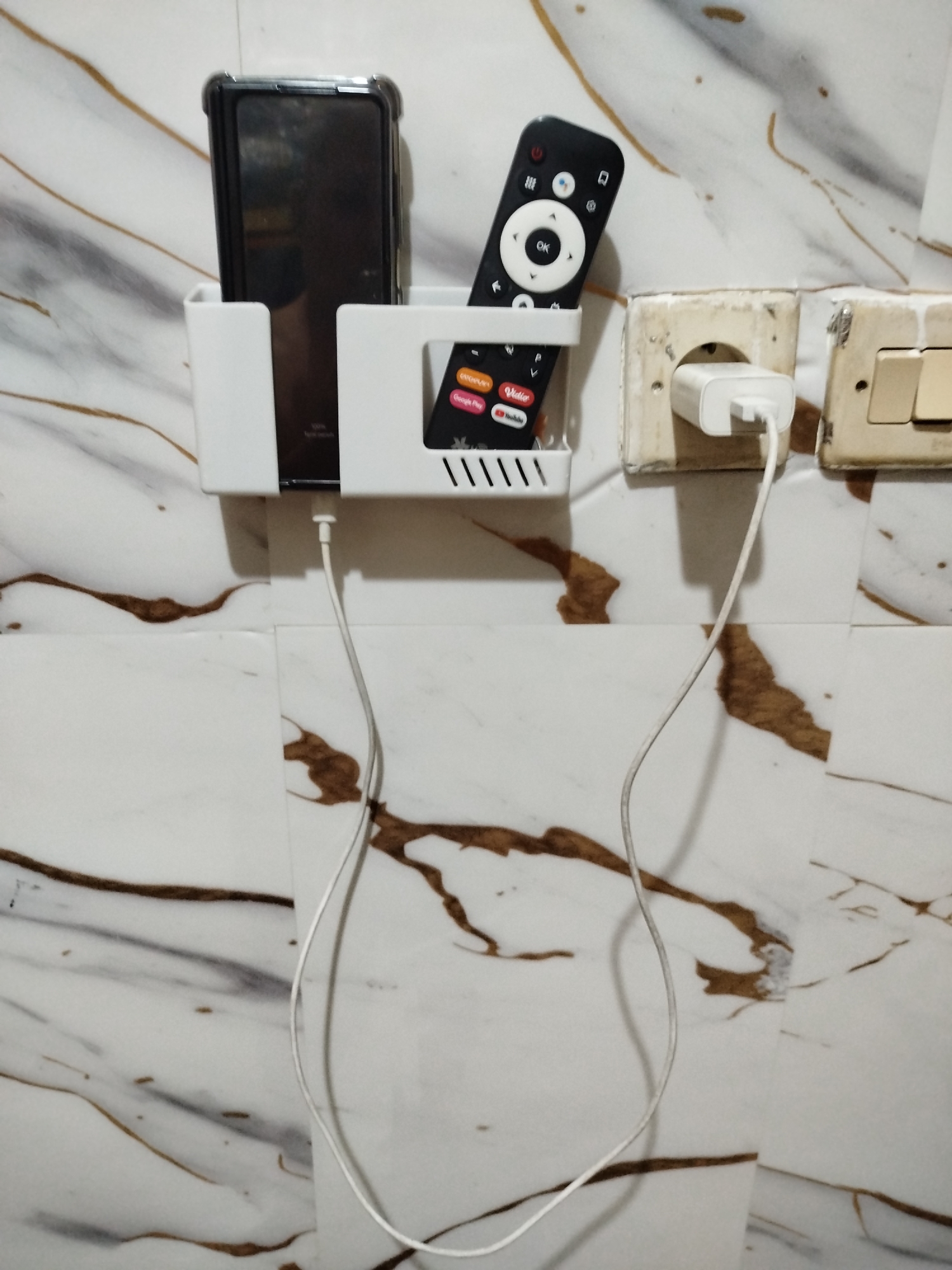 A white plastic organizer is mounted on a marble-patterned wall, holding a black smartphone and a remote control. The smartphone is in the left compartment, and the remote is in the right. A white charging cable connects the smartphone to a white power adapter plugged into a wall socket on the right. The wall has a white and brown marble pattern, and there is a light switch to the right of the power socket. The scene appears to be indoors, possibly in a bedroom or living area, in Bandungrejo, Indonesia.