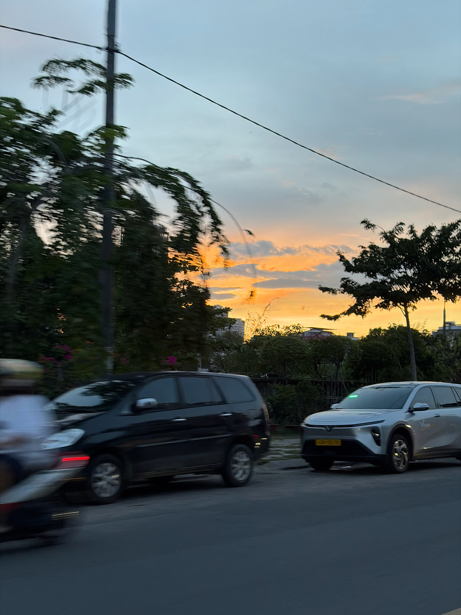 The image captures a street scene in Da Nang, Vietnam, during sunset. A silver SUV with a green light illuminated on its dashboard is parked on the right side of the road, facing forward. To its left, a black SUV is also parked. In the foreground on the far left, a blurry image of a motorcycle with a rider is captured in motion. The sky is filled with warm hues of orange and yellow as the sun sets, with clouds scattered across it. Trees and power lines line the street, creating a typical urban landscape. The overall impression is a fleeting glimpse of daily life against the backdrop of a beautiful sunset.