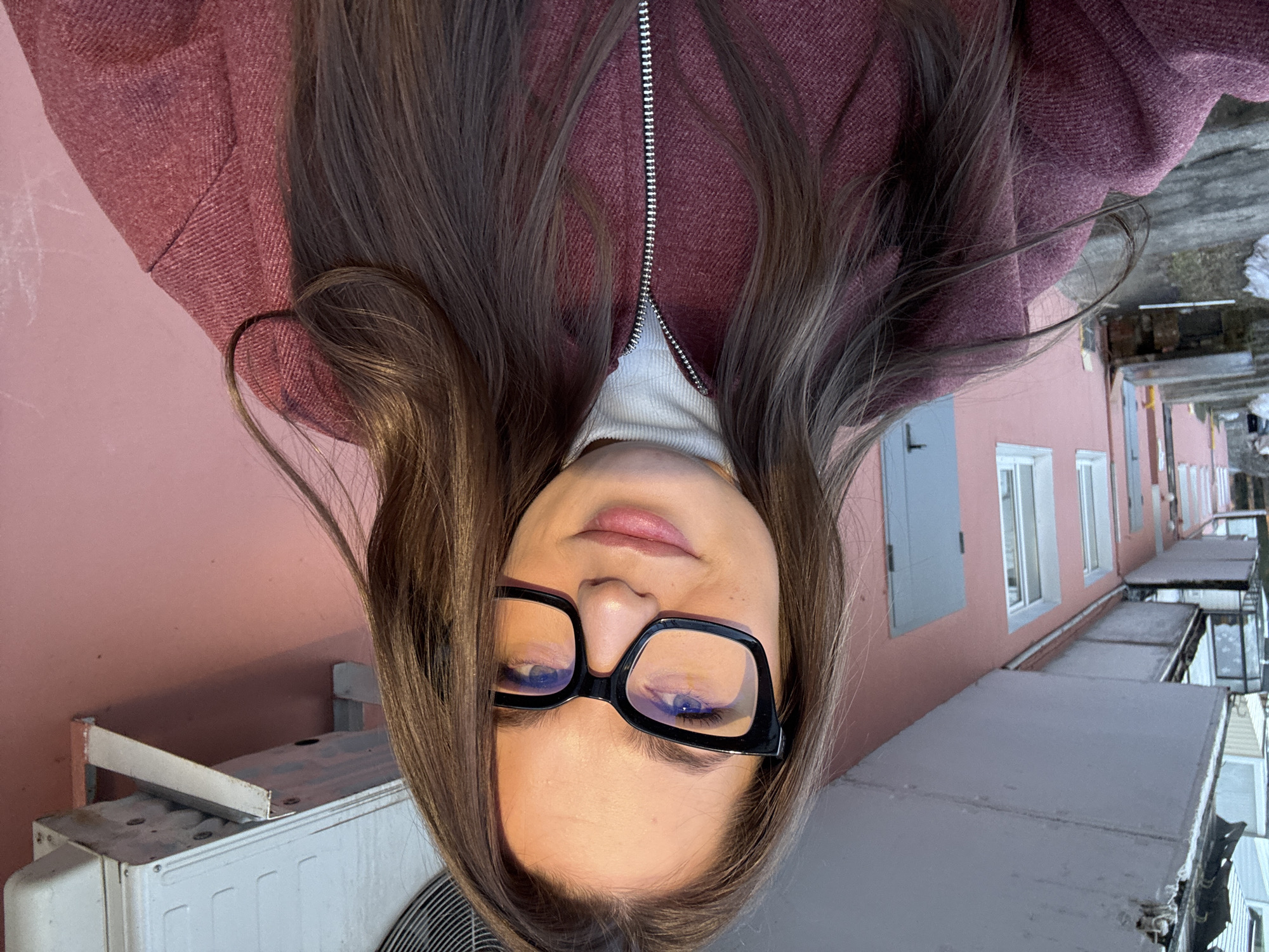 A person with long brown hair and glasses is shown in a selfie-style shot with the camera pointed upwards. The person is wearing a maroon jacket and a white turtleneck sweater. The background includes a pink building with grey window shutters and white-framed windows. A white air conditioning unit is also visible. The location is Chaikovsky, Russia.