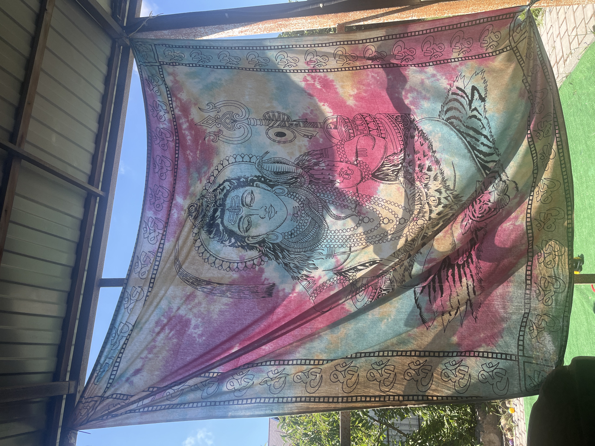 A vibrantly colored tapestry featuring an image of the Hindu deity Shiva is hanging outdoors. The tapestry has a tie-dye background in shades of pink, blue, and yellow, with a black outline drawing of Shiva in the center. Shiva is depicted with multiple arms, a serene expression, and intricate jewelry. The tapestry is bordered with a repeating pattern of the Om symbol. It appears to be hanging from a wooden structure with a metal roof, likely a gazebo or awning. In the background, there is greenery, a brick pathway, and a clear blue sky with some clouds. The scene is bathed in sunlight, casting shadows on the tapestry. The setting is outdoors, possibly in a residential backyard or a public outdoor space.