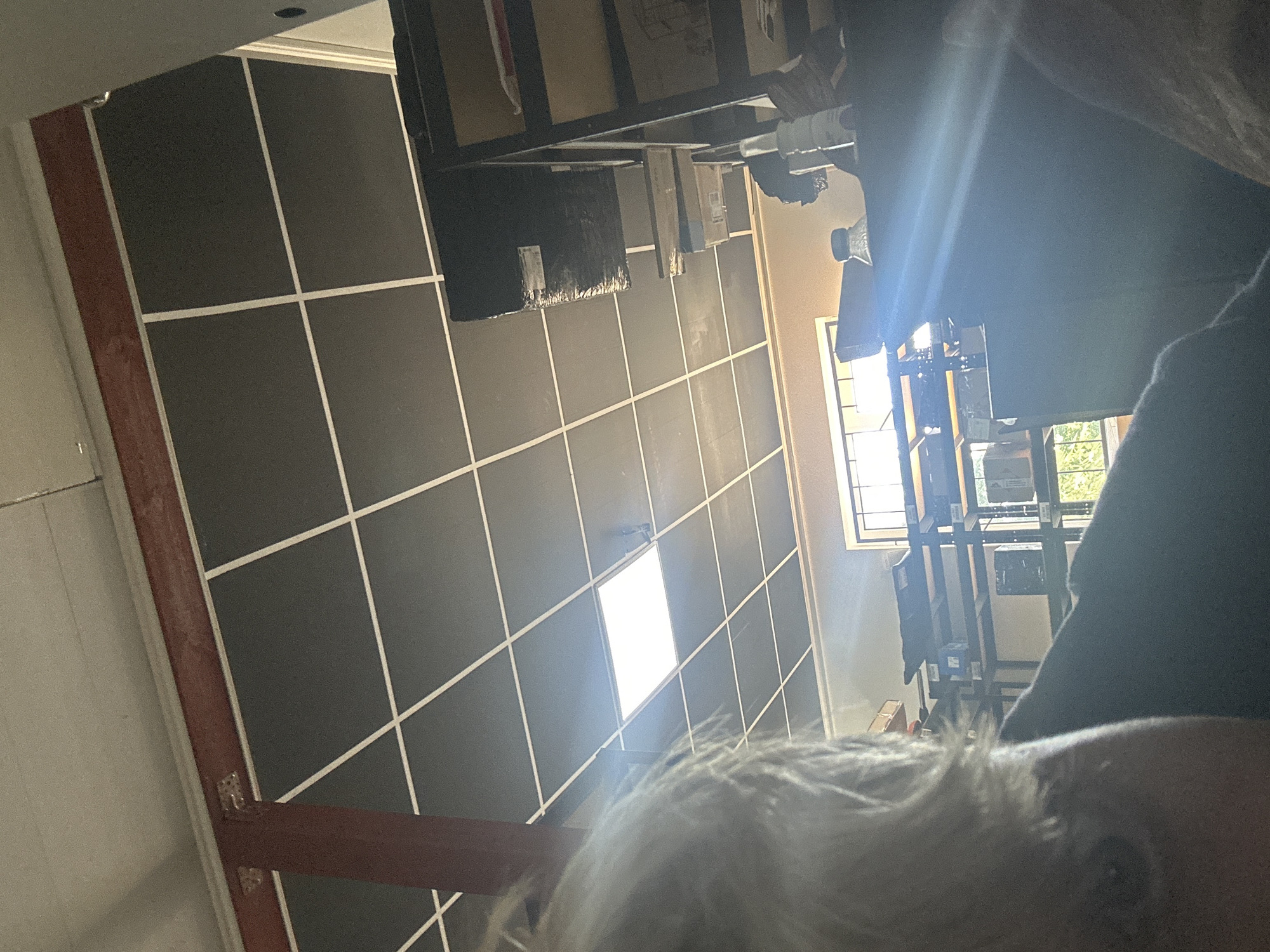 The image is a low-angle, slightly blurry shot looking up at a ceiling grid and a room beyond. The ceiling is made of dark square panels with white grid lines. A red beam is visible in the upper left, attached to the ceiling. A rectangular light fixture is illuminated in the center of the ceiling. Below the ceiling, the room appears to be a workshop or storage area with shelves and various objects, including boxes and what looks like a bottle. Sunlight streams into the room through a window with a grid pattern. In the foreground, a person's head and shoulder are visible, out of focus, with light-colored hair. The specific location in Khmilnyk, Ukraine cannot be determined from the image.