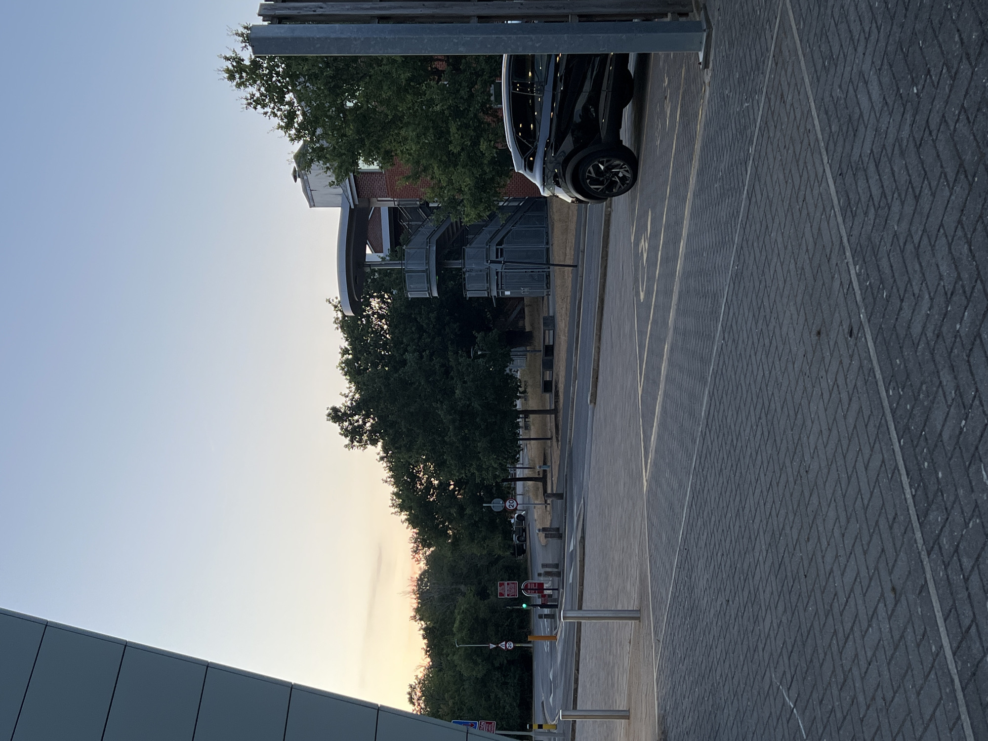The image depicts an outdoor scene, likely a university campus or a business park, in Guildford, United Kingdom, during what appears to be early evening or late afternoon due to the soft lighting and the warm hues in the sky. In the foreground, a paved area with a herringbone brick pattern is visible, marked with yellow lines indicating parking spaces. A black SUV is parked in one of these spaces, partially obscured by a wooden post.
To the left, a portion of a modern building with a grey, angled facade is present. Beyond the paved area and across a road, a cluster of mature trees forms a green backdrop. In the middle ground, a building with red brick and a grey external staircase with a circular platform at the top is visible amongst the trees. There are also streetlights and road signs, including speed limit signs indicating 20. Traffic lights are visible, showing a green light. A few benches are also present in the background. The sky is a clear gradient of pale blue and yellow, suggesting the sun is low on the horizon. No people are clearly visible in the image.