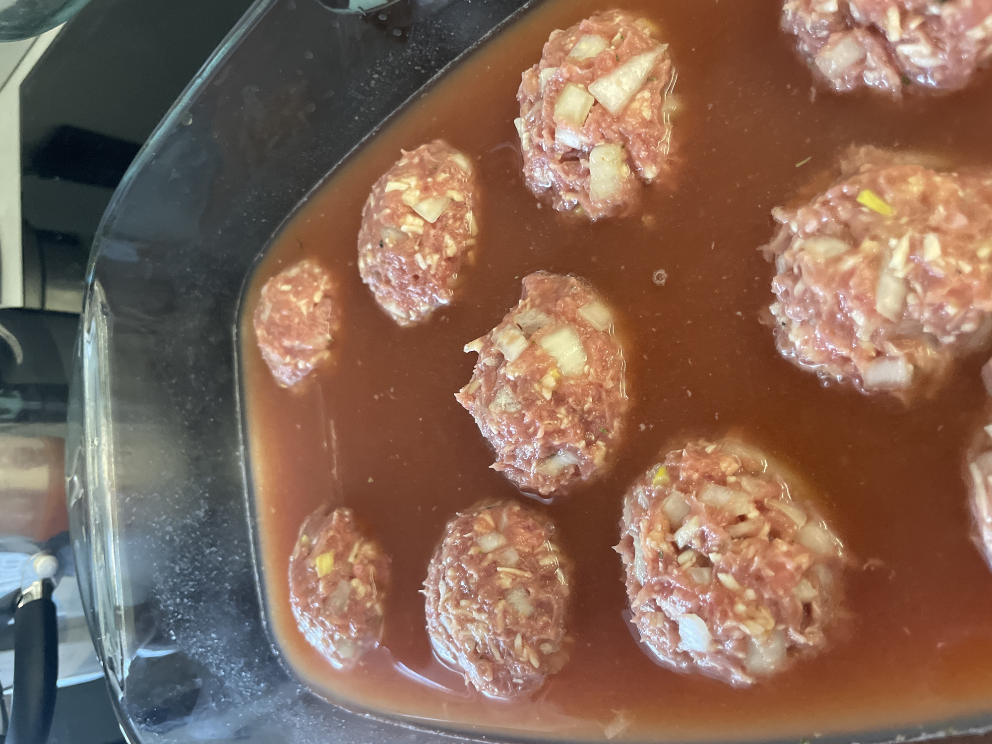 The image shows a clear glass baking dish containing approximately nine raw meatballs submerged in a reddish-brown sauce. The meatballs appear to be made of ground meat mixed with diced white onions. The sauce level is such that it nearly covers the meatballs. The scene is likely indoors, possibly in a kitchen. There is no information to suggest a location in Izhevsk, Russia or any other specific location.