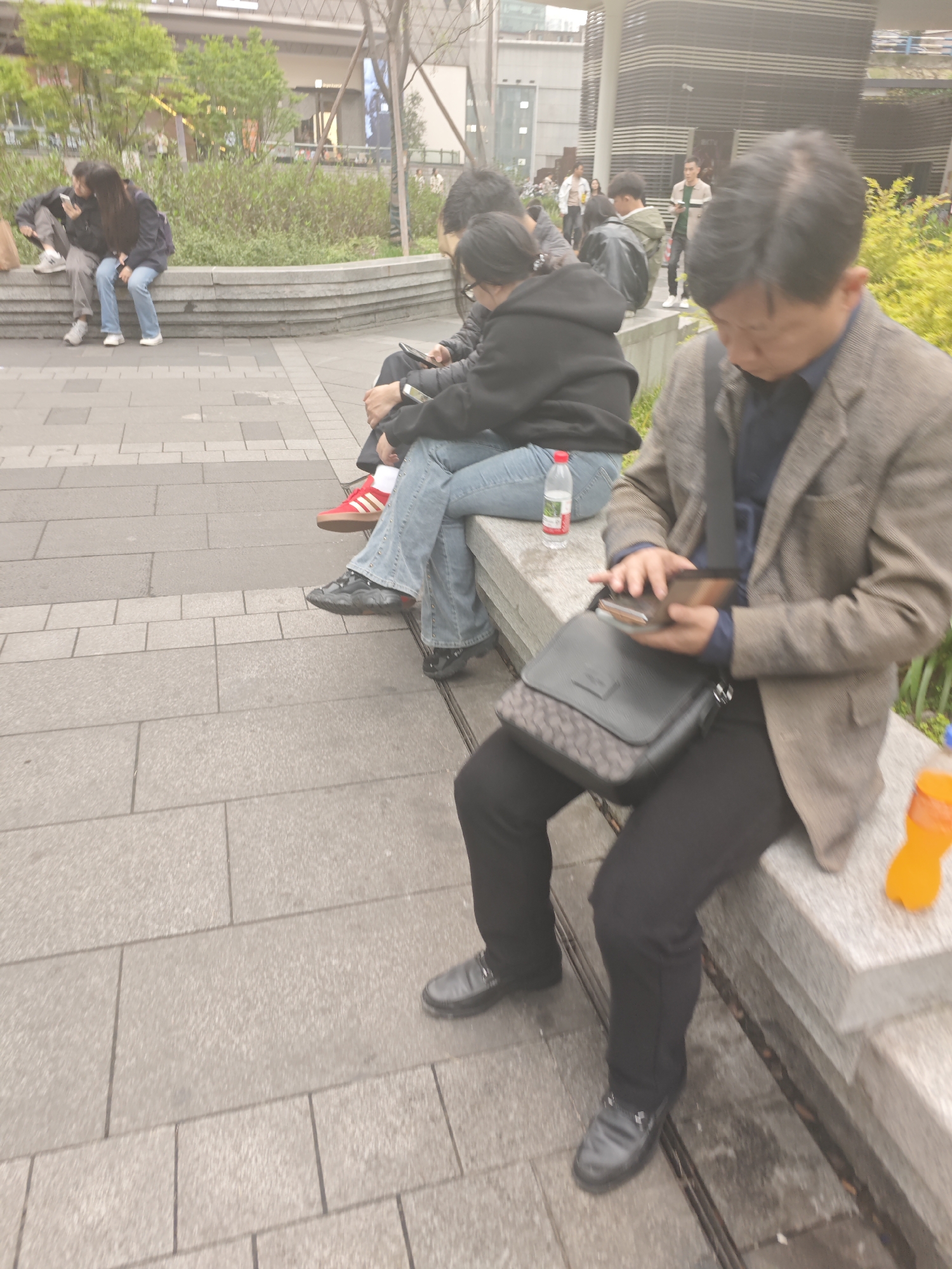 The image depicts a public outdoor scene in what appears to be an urban setting. Several people are present, some sitting on a low concrete wall and others walking in the background. A man in the foreground is seated, wearing a brown jacket and black pants, and looking at a mobile phone in his hands. He has a black shoulder bag at his side. Other individuals sit to the left, one wearing a black hoodie and jeans, and another in red sneakers. A plastic water bottle and orange soda are near the sitting man. The background includes greenery, buildings, and other pedestrians. It is in Liangjiang, China.