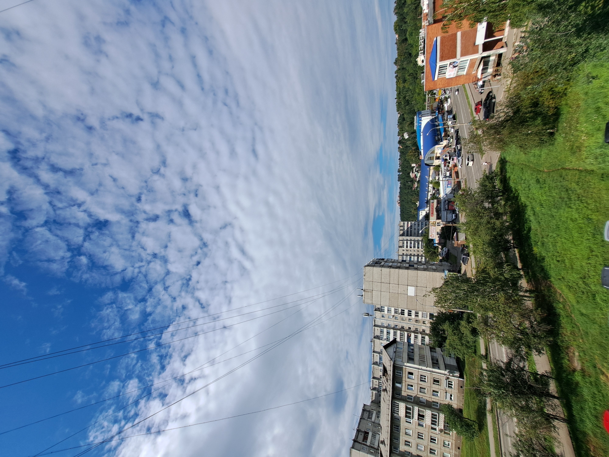 The image is an aerial view of a city street in Irkutsk, Russia, on a partly cloudy day. The foreground features a lush green grassy area and trees. A paved road runs horizontally across the middle ground, with several cars driving on it. On the left side of the road, there are multi-story apartment buildings, typical of Soviet-era architecture, with balconies and numerous windows. On the right side of the road, there are commercial buildings, one of which is made of red brick and has blue accents. In the distance, a densely wooded hill can be seen, with some houses nestled among the trees. Overhead, a network of power lines crisscrosses the sky, which is a vibrant blue with scattered white clouds. The scene depicts a typical urban landscape with a mix of residential and commercial structures, set against a natural backdrop.