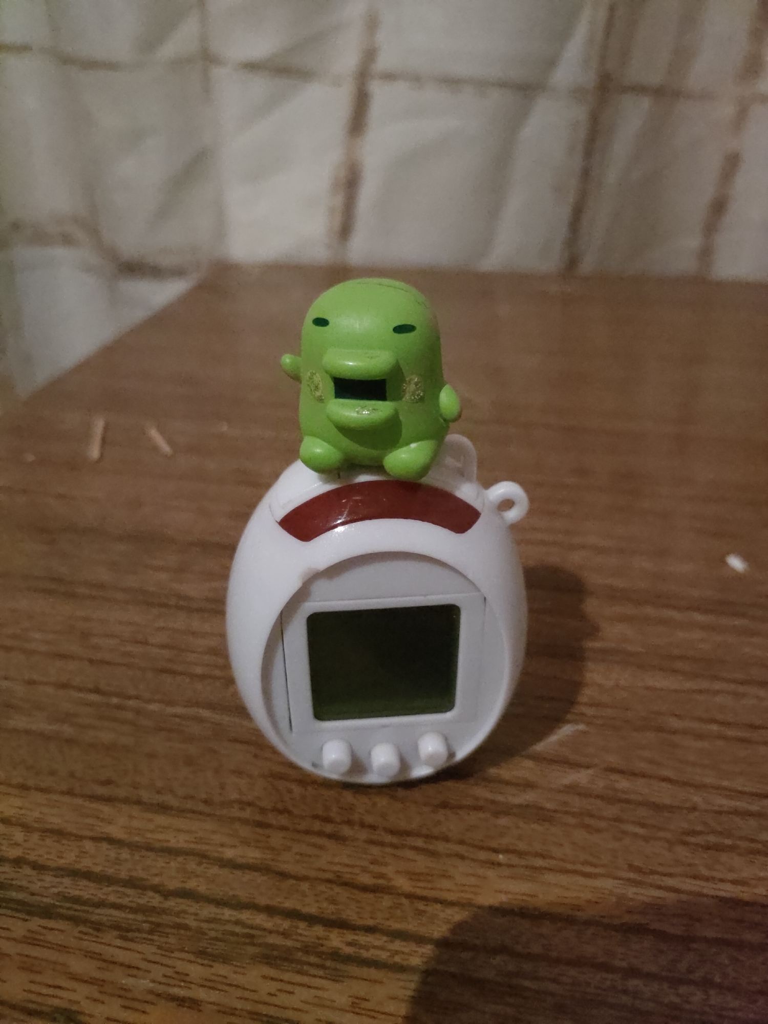 A small, green, cartoonish character with a wide mouth and simple eyes sits atop a white digital pet toy. The toy has a red accent above its screen and three white buttons below it. The objects are placed on a wooden surface, possibly a table, with a blurred background of white curtains or fabric. The scene is lit from the front, casting a subtle shadow of the objects onto the wooden surface. The photograph appears to be taken indoors. There is no visible indication of the location being in Дзешчанскі сельскі Савет, Belarus.