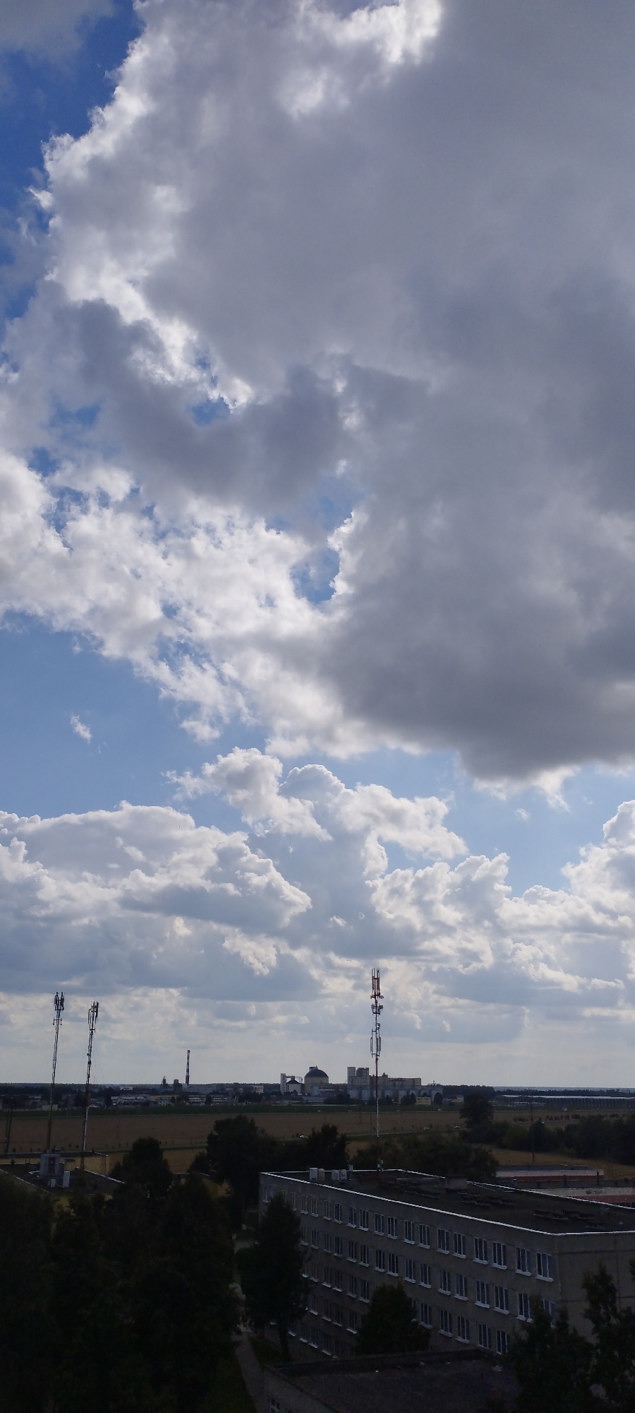 The image captures a wide, open landscape under a dramatic, cloudy sky. The sky is filled with a mix of bright white and darker gray cumulus clouds, interspersed with patches of clear blue sky, suggesting a dynamic weather pattern, likely during the daytime.
In the mid-ground, a cluster of industrial buildings with domed roofs and tall, slender smokestacks are visible, hinting at a manufacturing or power generation facility. A few communication towers, one prominently featuring red and white sections, punctuate the horizon, indicating a developed area. The land between the foreground and the industrial complex appears to be agricultural or undeveloped fields, with some scattered trees.
The foreground of the image is dominated by the tops of buildings. On the right, a long, multi-story building with numerous windows runs horizontally across the frame. Dark trees obscure parts of the landscape and other structures.
The overall scene suggests an industrial or semi-urban fringe area. The lighting implies it is daytime, likely late morning or afternoon, given the brightness of the sun filtering through the clouds. There are no people visible, and the focus is on the landscape and structures. No text is discernible in the image.
