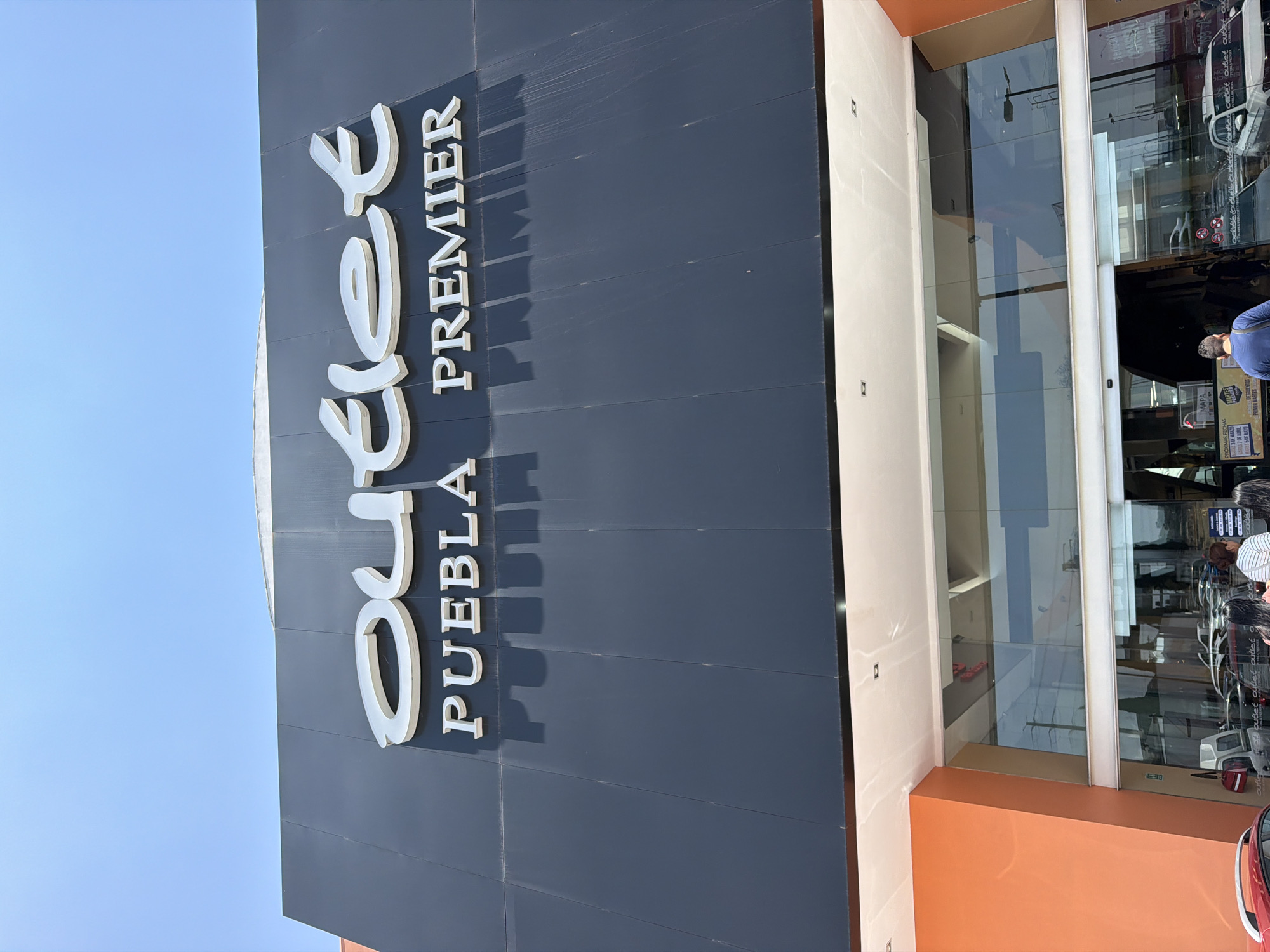 Here's a detailed description of the image:
**Overall Scene:** The image captures the exterior of the "Outlet Puebla Premier," a shopping outlet located in San Francisco Ocotlán (Ocotlán), Mexico. The perspective is from a low angle, looking upwards at the building.
**Building Facade:** The building features a modern design. A prominent dark gray wall displays the outlet's name in large, vertically oriented white letters with shadows. To the right of the wall is a glass-fronted entrance, partially reflecting the surroundings. The entrance is framed by light beige walls and an orange accent at the bottom.
**Signage:** The white "Outlet Puebla Premier" sign is the focal point of the image. The text is stacked vertically, with "Outlet" stylized in cursive.
**People:** Although not the main subject, glimpses of people are visible through the glass entrance. Some are facing into the building, while others are slightly blurred, suggesting movement.
**Objects:** Inside the glass facade of the store entrance there are a map and a list of upcoming events and promotions.
**Environment:** A clear blue sky is visible above the building, suggesting a sunny day.