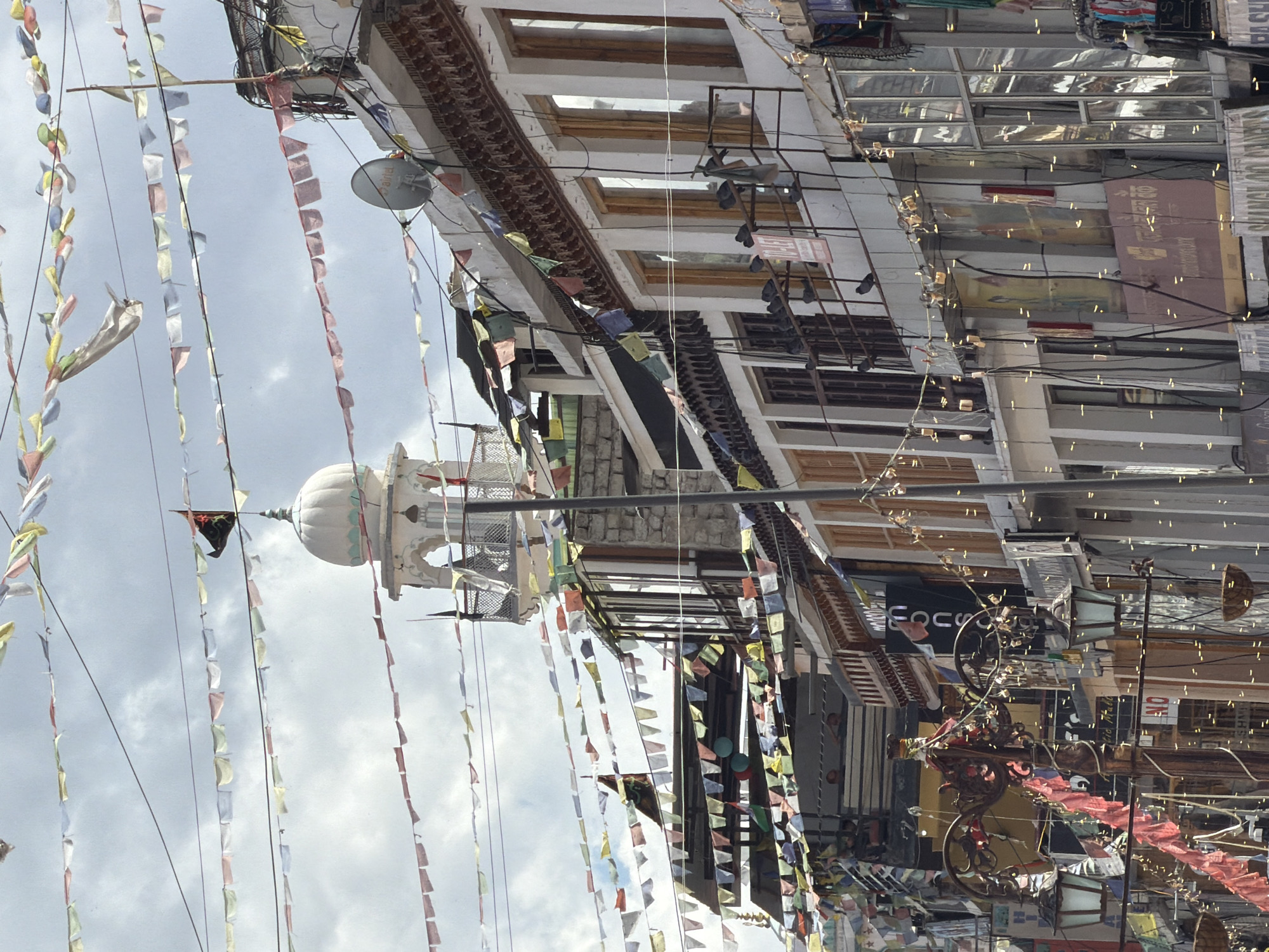 The image captures a street scene in Leh, India, likely along a main thoroughfare. The foreground features two ornate lampposts adorned with strings of small, yellow lights, and a few hanging baskets. Several buildings line the street, displaying a mix of architectural styles. Prayer flags, in various colors, are strung across the sky, creating a festive atmosphere. A prominent white domed structure with minarets stands out against the cloudy sky, suggesting a religious or historical building. The buildings themselves have multiple stories with wooden-framed windows and balconies. Signs and advertisements are visible on the storefronts, indicating commercial activity. Pigeons are perched on railings and ledges of the buildings. The overall impression is of a bustling, culturally rich urban environment in a mountainous region.