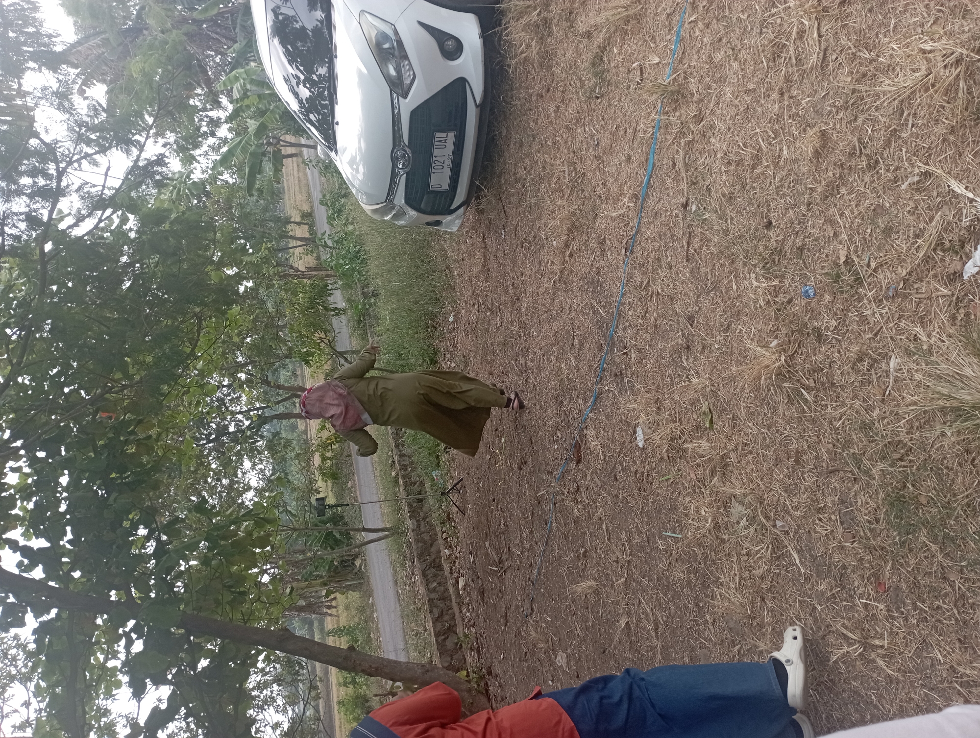 The image depicts an outdoor scene with dry, grassy ground and sparse trees in the background. A white car, with a license plate reading "D 1021 UAL 10-27", is parked on the right side of the frame. In the center, a person wearing a green abaya and a pink headscarf is seen from behind, walking away from the camera with their arms outstretched. To the left, a person's legs are visible, wearing blue jeans and white clogs. A tripod with a camera or phone is set up in the middle ground, near a low stone wall. The environment appears to be rural, possibly a roadside or open field, with evidence of agricultural land in the distance. The overall lighting suggests an overcast or diffused daylight. While the image doesn't provide explicit visual cues for Purwakarta, Indonesia, the license plate format is consistent with Indonesian vehicles.