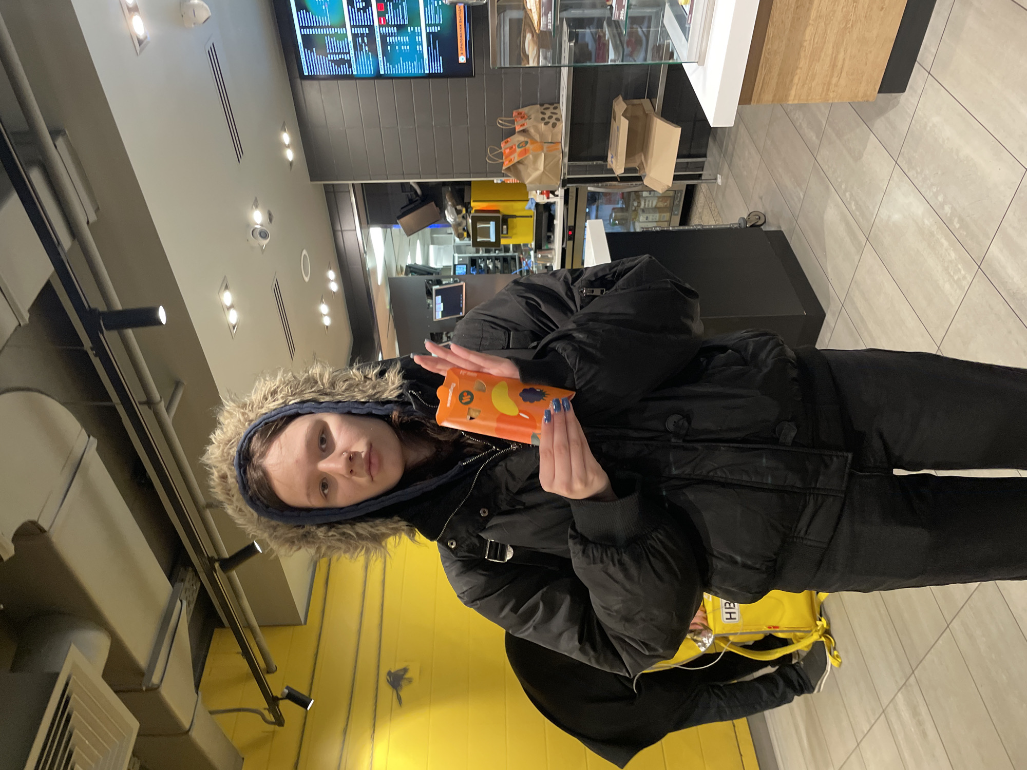 The image depicts a young person standing in what appears to be a fast-food restaurant or food court in Saint Petersburg, Russia. The individual, likely female, wears a black winter jacket with a fur-lined hood pulled up around her head, suggesting cold weather outside. She is holding a small, orange rectangular object, possibly a phone in a unique case or a small food box, featuring cartoon-like graphics. Her fingernails are painted blue.
The background shows a typical quick-service establishment setting. Behind her, a bright yellow wall features a faint, dark silhouette of an object, possibly a bird or a logo. To her right, a service counter with display cases holds various items, and a few brown paper takeaway bags are visible. Above the counter, digital menu boards display text in Cyrillic script; one clearly visible phrase reads "ВРЕМЕННО ВЫХОДИТ" (VREMENNO VYKHODIT), which translates to "TEMPORARILY OUT" or "TEMPORARILY UNAVAILABLE." The ceiling is equipped with multiple recessed lights, ventilation grates, and a security camera. The floor is tiled in a light gray pattern. A yellow bag or backpack with the letters "HB" partially visible hangs near her side. The overall scene captures a casual moment indoors.