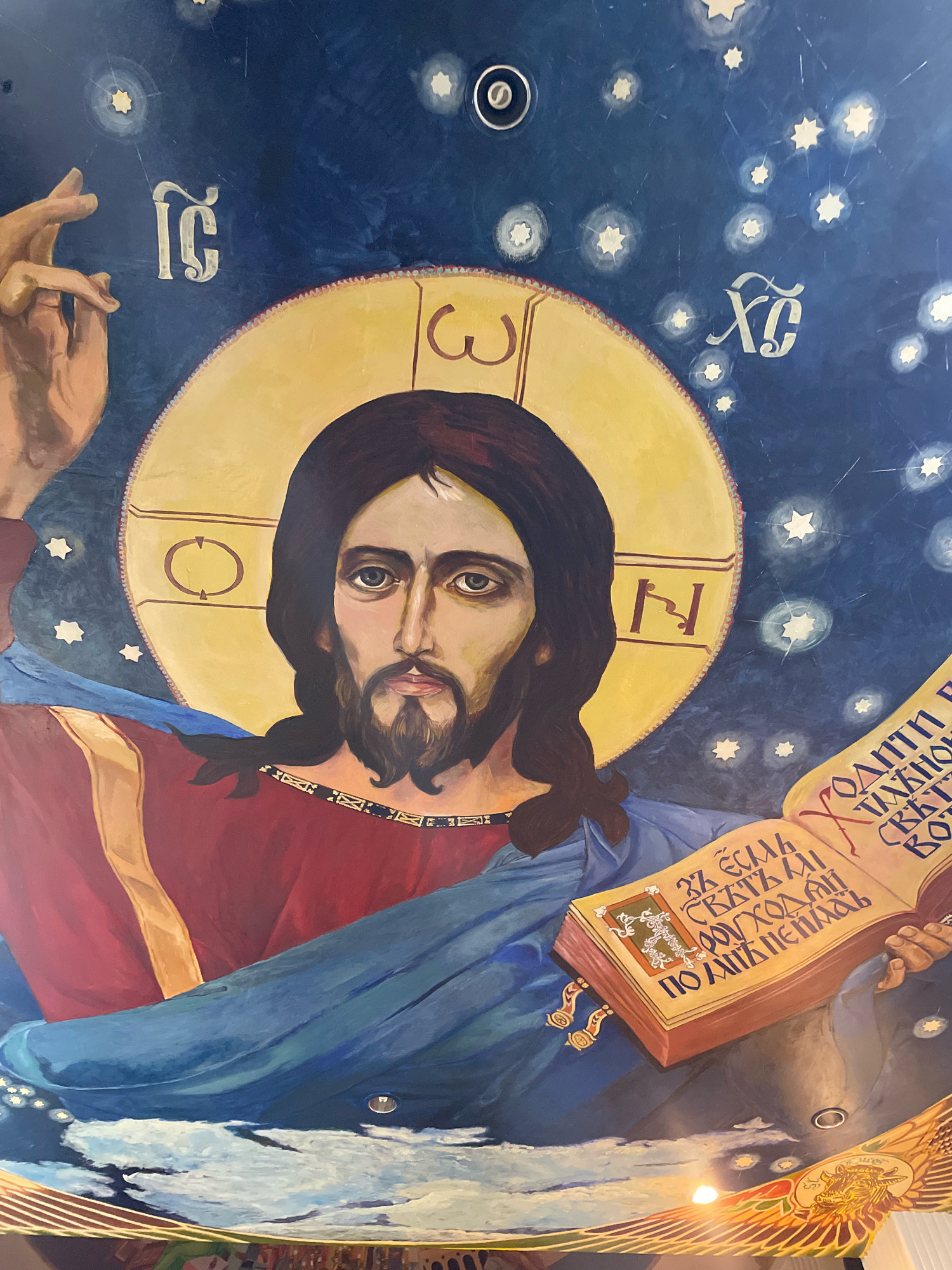 The image is a close-up, low-angle photograph of a mural depicting Jesus Christ on a dark blue background with stars. Jesus is centrally positioned, facing forward with dark hair, a beard, and piercing eyes. He is wearing a red garment and a blue robe draped over his shoulder. His left hand is raised, with fingers extended, while his right hand holds an open book with text and illustrations. The book is adorned with golden clasps. There are Greek letters visible on the mural, specifically "IC" and "XC" to the left and right of Jesus's halo, respectively, which are abbreviations for Jesus Christ. Additionally, there are other Cyrillic or Greek-like symbols within the halo and on the book. The mural appears to be part of a larger artwork, with hints of other decorative elements at the bottom edge. The lighting suggests the photograph was taken indoors, potentially in a church or religious establishment, and there is a circular light fixture visible on the ceiling above Jesus's halo.
This mural is located at the Sanctuary of Truth in Pattaya City, Thailand. The Sanctuary of Truth is a colossal wooden structure that is entirely constructed from wood, employing traditional Thai craftsmanship and philosophy, and is dedicated to the ancient knowledge and philosophies of this region, along with inspiring philosophical teachings.