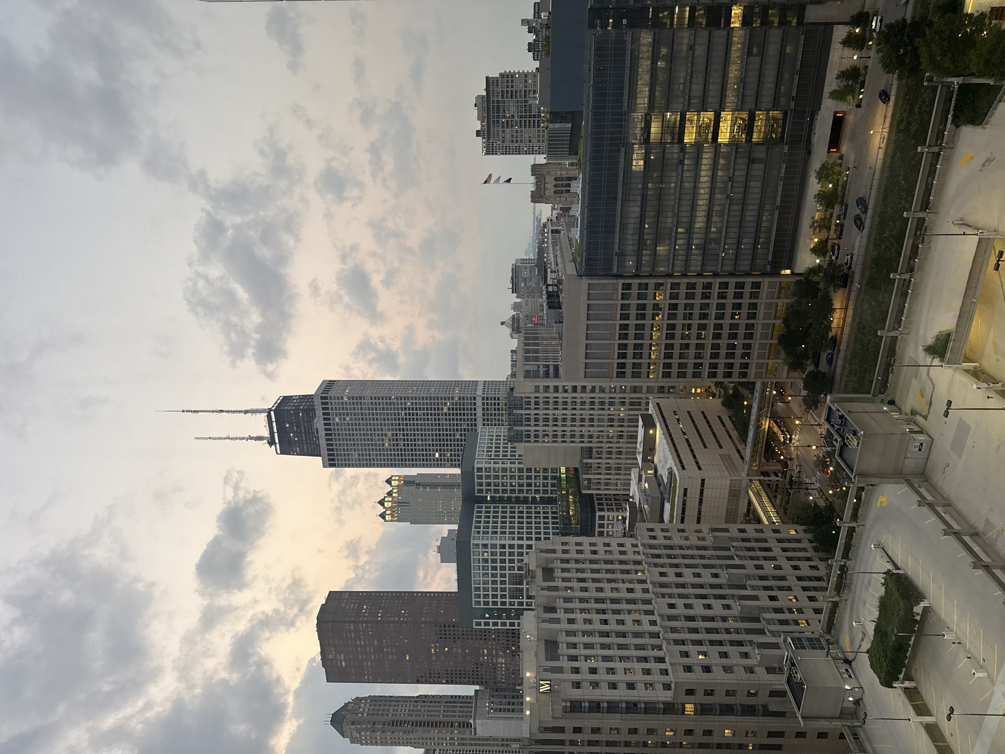 This is a daytime, eye-level shot of the Chicago skyline under a cloudy sky. The image captures a dense collection of skyscrapers, with the Willis Tower prominently visible in the background, distinguished by its height and twin antenna spires. The architecture varies from the modernist glass and steel structures to older, more solid buildings. The foreground features a multi-level parking garage, with concrete ramps, yellow lane markings, and some greenery on one of the levels. Several cars are visible on the street level below the parking garage, and streetlights are also present. The overall impression is that of an urban environment, likely during the late afternoon or early evening, given the overcast conditions and the lights beginning to illuminate some of the buildings.