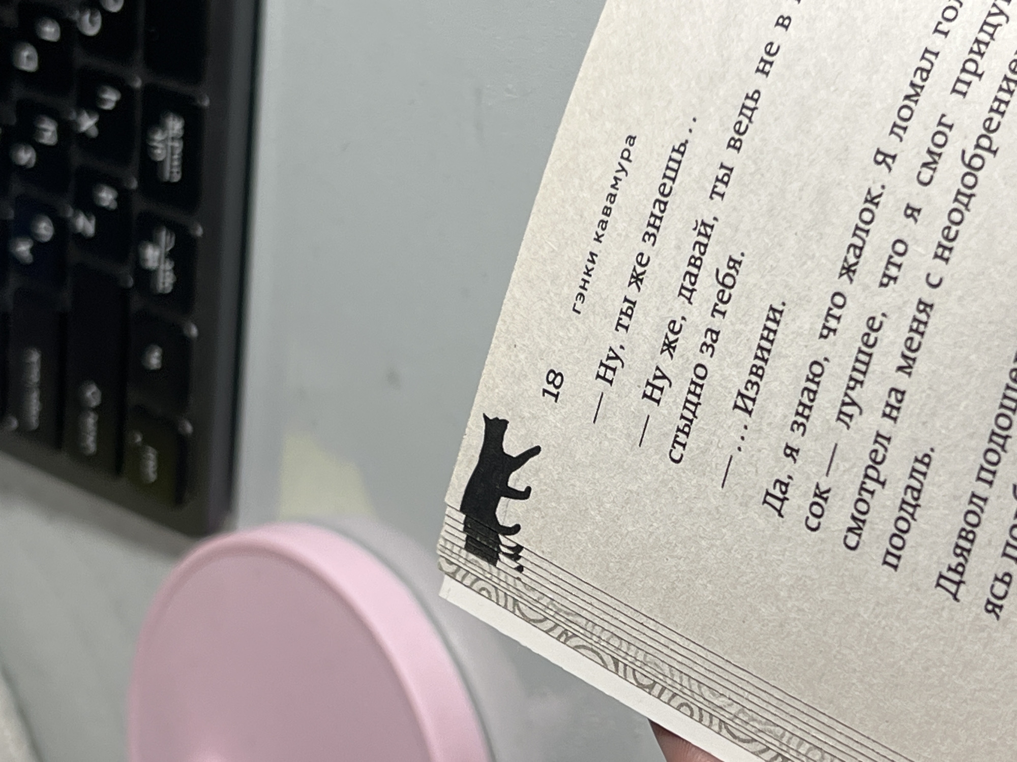 A close-up overhead shot shows an open book lying on a desk. The book has a black cat illustration on the left side of the page, as if it is a bookmark. The text in the book is in Russian. To the left of the book, a pink object, possibly a mousepad or a coaster, is visible. In the background, a black keyboard is partially visible. The scene appears to be indoors, with the lighting suggesting it is daytime. No people are visible in the frame.