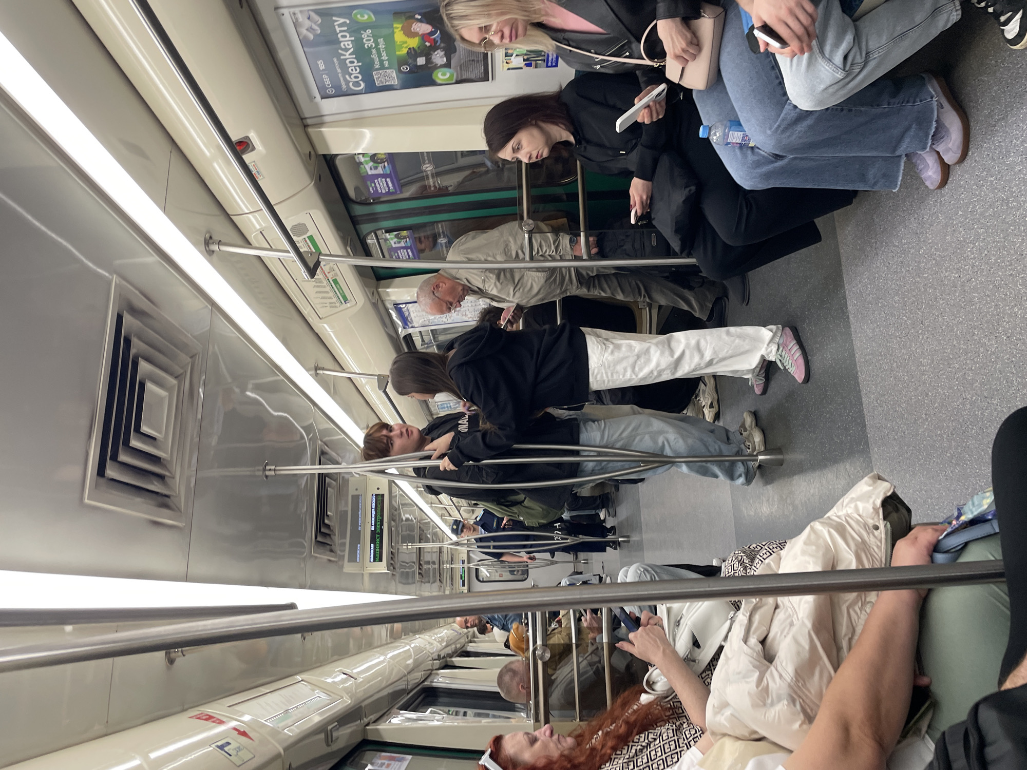The image depicts the interior of a Saint Petersburg Metro train car. Several passengers are visible, both seated and standing. In the foreground, a woman with red hair is seated on the left, looking at her phone. To her right, a man with a shaved head is also seated. Further back, two young people are engaged in a conversation, standing in the aisle. A uniformed metro worker is visible walking down the aisle. Several other passengers are seated or standing throughout the car, with some looking at their phones. The environment is typical of a metro train, with metal poles for support, illuminated lighting strips on the ceiling, and advertisements displayed on the walls. The floor is a grey, speckled material. The overall impression is that of a busy commuter train during a regular operational period.