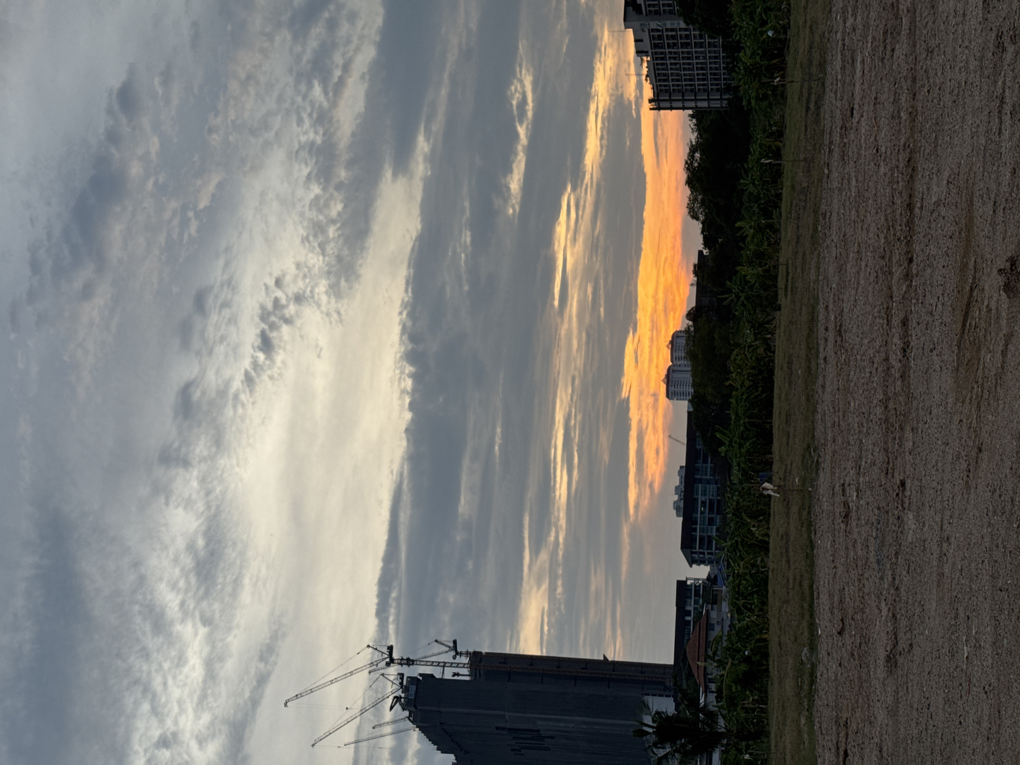 The image shows an outdoor scene in Pattaya City, Thailand. In the foreground is a dirt/gravel ground area transitioning into a grassy field. To the left, there is a tall building under construction, with several cranes visible. Beyond the construction site are residential buildings and trees. Above the buildings, the sky presents a blend of gray and white clouds, with a patch of orange color towards the center indicating either sunrise or sunset. The buildings, vegetation, and sky together create a somewhat urban landscape.