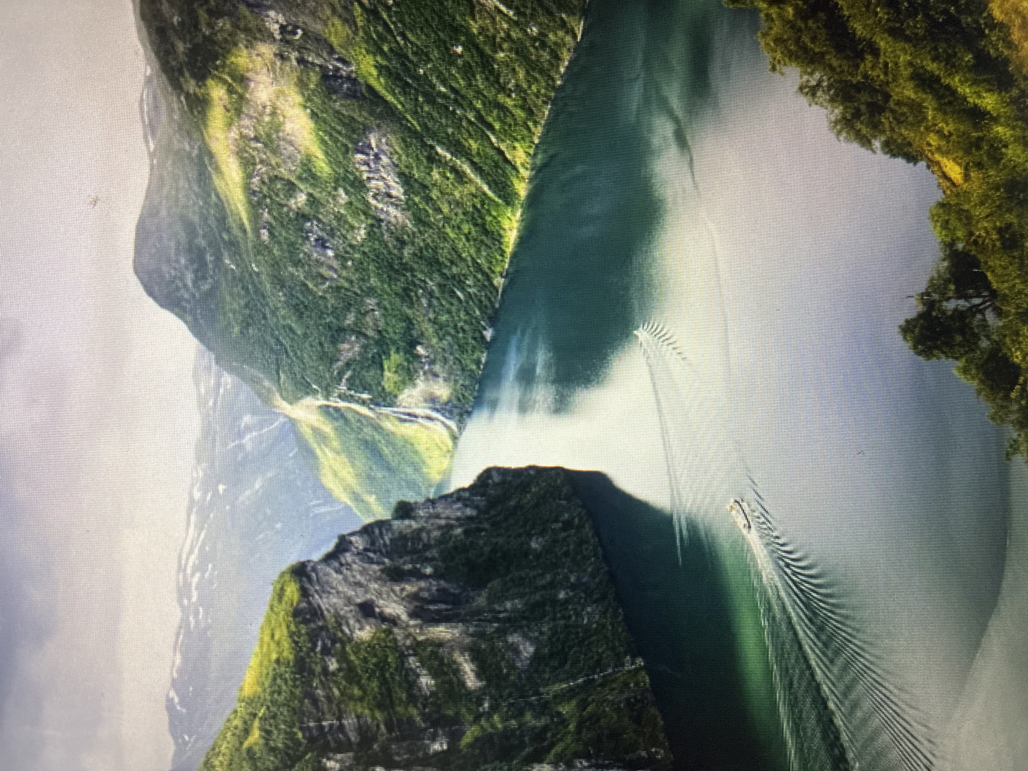 The main image is an upward-tilted photograph of a computer screen, displaying a majestic landscape scene. The displayed image itself is rotated approximately 90 degrees counter-clockwise.
**Subjects:** The scene features a deep, narrow body of water, likely a river or reservoir, flanked by incredibly steep and tall mountains. These mountains are densely covered in vibrant green vegetation and trees, with some visible rocky outcrops. A small, light-colored boat is cruising across the water. The upper left portion of the image shows a bright sky or distant, hazy mountainous terrain.
**Activity or event taking place:** A boat is moving across the water, creating a prominent, V-shaped wake that trails behind it, indicating movement.
**Notable details:** The time of day appears to be bright daylight, with the sun illuminating the scene and creating areas of both light and shadow on the water and mountain slopes. The weather seems clear or partly cloudy, offering good visibility of the distant features. The overall impression is one of grandeur and natural beauty. Given the location context of Asenovgrad, Bulgaria, this landscape likely depicts a river gorge or a reservoir within the mountainous Rhodope region. A distinct grid pattern across the entire image confirms that it is a photograph taken of a digital screen.
**Visible text:** No visible text is present in the image.