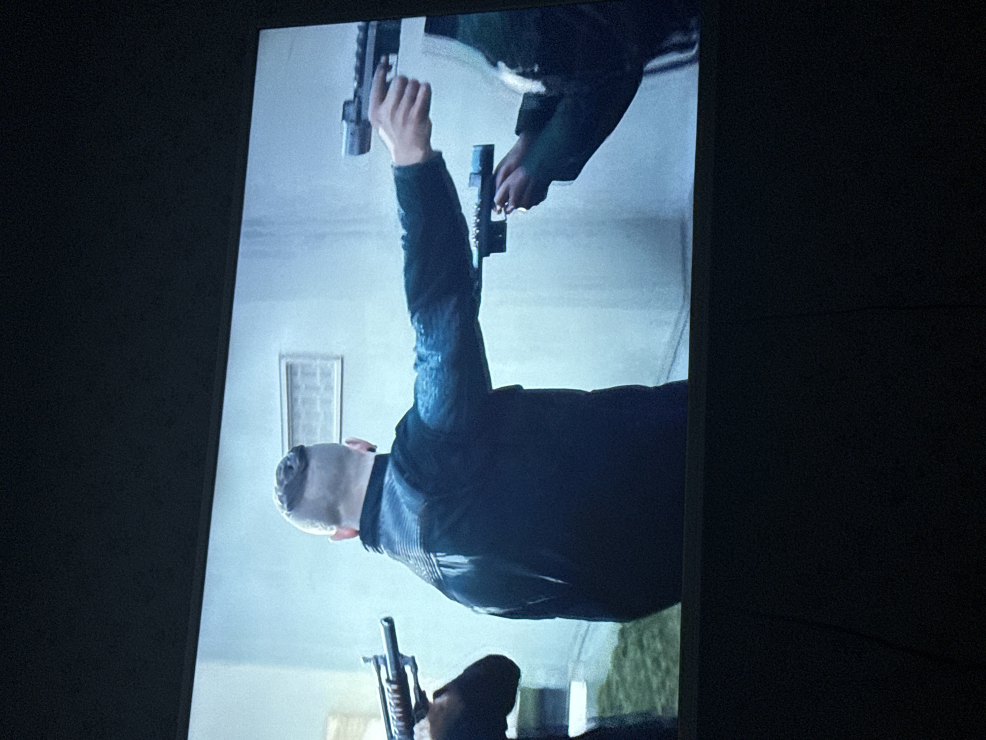 The image shows a still from a video, with the screen rotated counter-clockwise. Two individuals are visible holding firearms. One individual is bald, wearing a dark jacket with a ribbed texture on the sleeves and back. They have their arm raised, holding a pistol upwards. Above them, another person is holding a pistol downwards. A third firearm is visible near the bottom edge of the screen. The environment appears to be indoors, possibly a room. No information indicates a connection to Mytishchi, Russia.