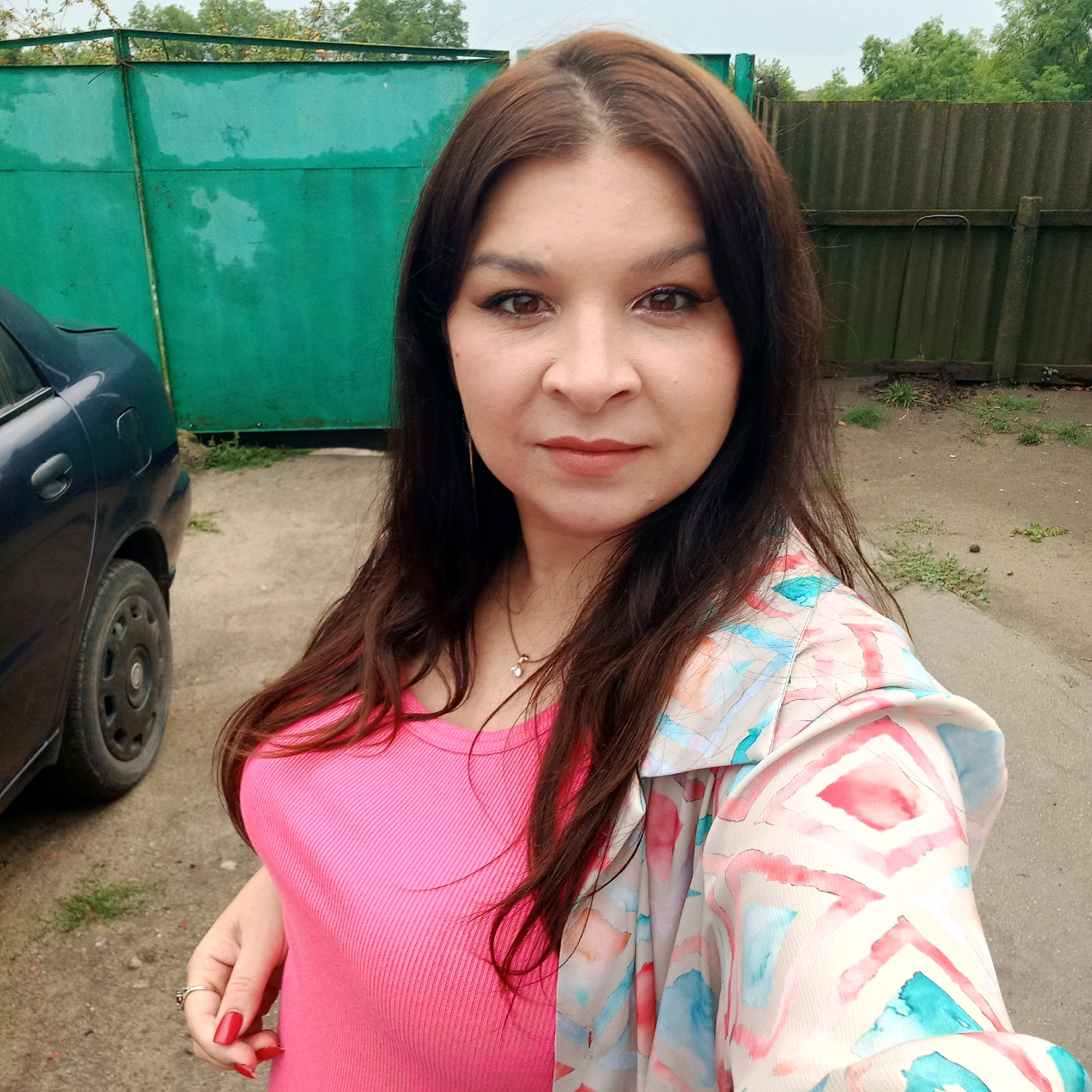 A woman with long brown hair and light skin takes a selfie outdoors. She is wearing a bright pink ribbed tank top and a light-colored jacket with a watercolor-like pattern of pink, blue, and coral diamond shapes. She has on makeup, including eyeliner and lipstick, and is wearing a delicate necklace with a small pendant and a ring on her left hand. Her fingernails are painted red.
She is standing in what appears to be a residential yard or driveway. To her left, the rear of a dark blue car is visible, with its rear wheel and part of the fender showing. Behind her is a tall, teal-colored metal fence. Further in the background, to her right, is a weathered, dark grey wooden fence, and beyond that, trees and foliage are visible under an overcast sky. The ground is a mix of gravel and dirt, with some patches of sparse grass. The location is identified as Klyntsi, Ukraine.