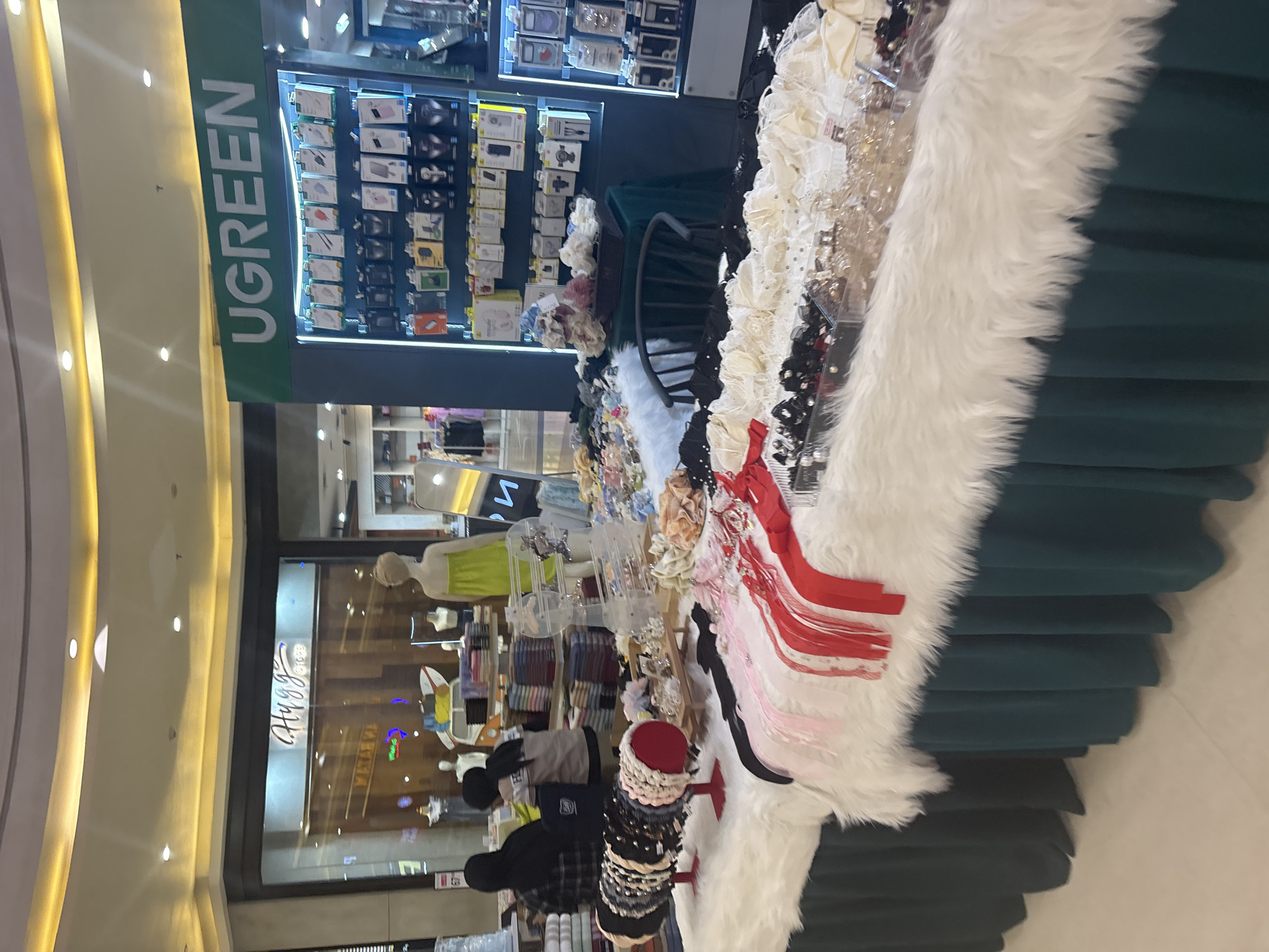 This image depicts an indoor market stall, likely within a shopping mall. The stall displays a variety of hair accessories, including headbands, hair clips, and ribbons, arranged on tables covered with white faux fur. Behind the accessory stall, a store with the brand name "UGREEN" is visible, showcasing electronic accessories like phone cases and chargers on illuminated shelves. In the background, mannequins dressed in clothing are displayed in a shop window with the brand name "Hugg." Two individuals are partially visible in the foreground, one wearing a black hijab and the other a grey and white top, appearing to be customers or vendors. The overall environment suggests a commercial setting within a mall in North Jakarta, Indonesia, characterized by bright lighting and a multitude of retail displays.