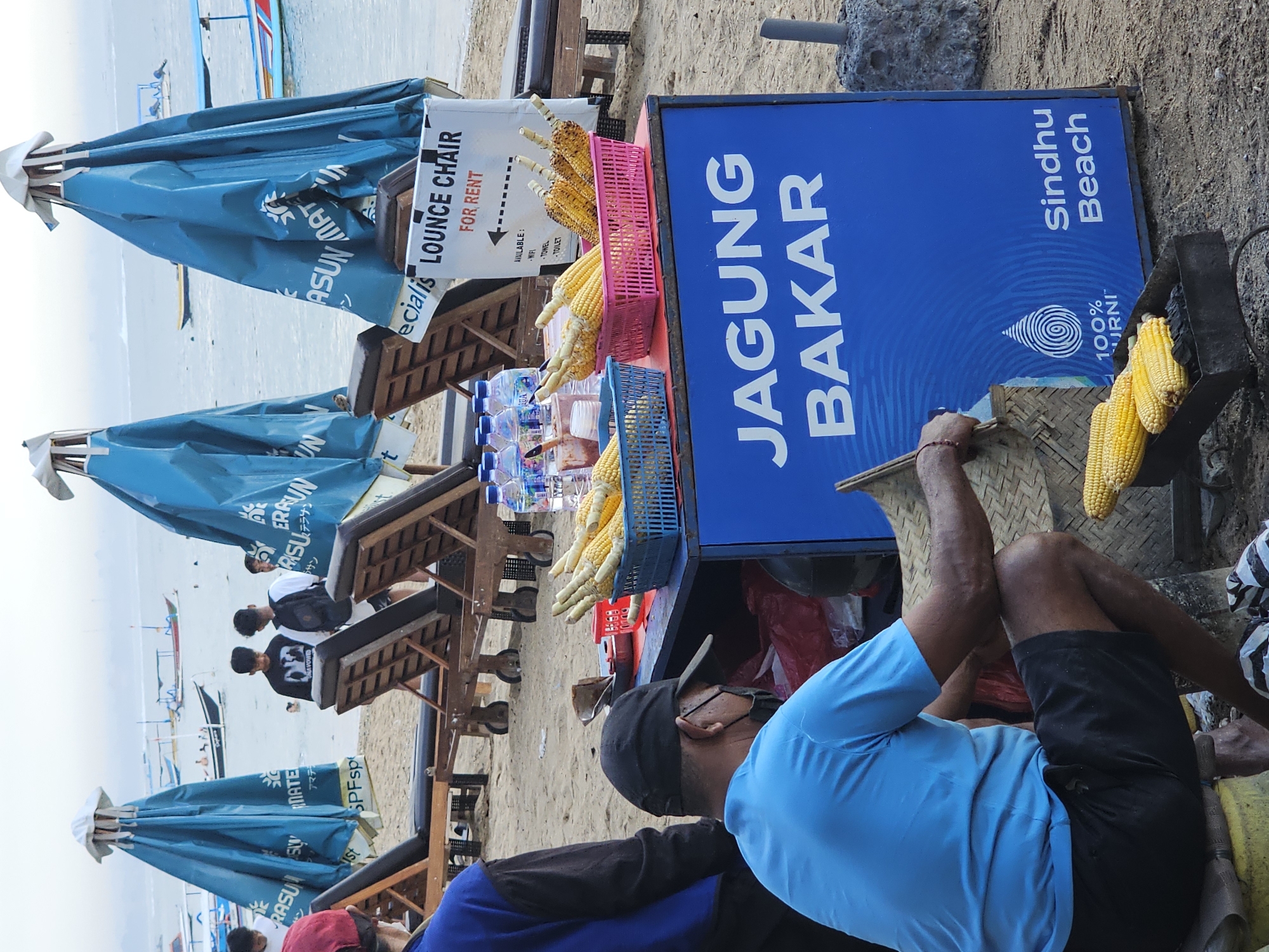 A man, wearing a blue t-shirt and black shorts, is crouching on a beach in Denpasar, Indonesia, grilling corn. He is wearing a black baseball cap and a face mask. In front of him is a blue cart with the words "JAGUNG BAKAR" (grilled corn) and "Sindhu Beach" written on it. There are baskets of corn on the cart and some corn is on a grill in front of the cart. In the background, there are beach chairs and blue umbrellas. The ocean is visible in the background with several boats on it. The sky is overcast.